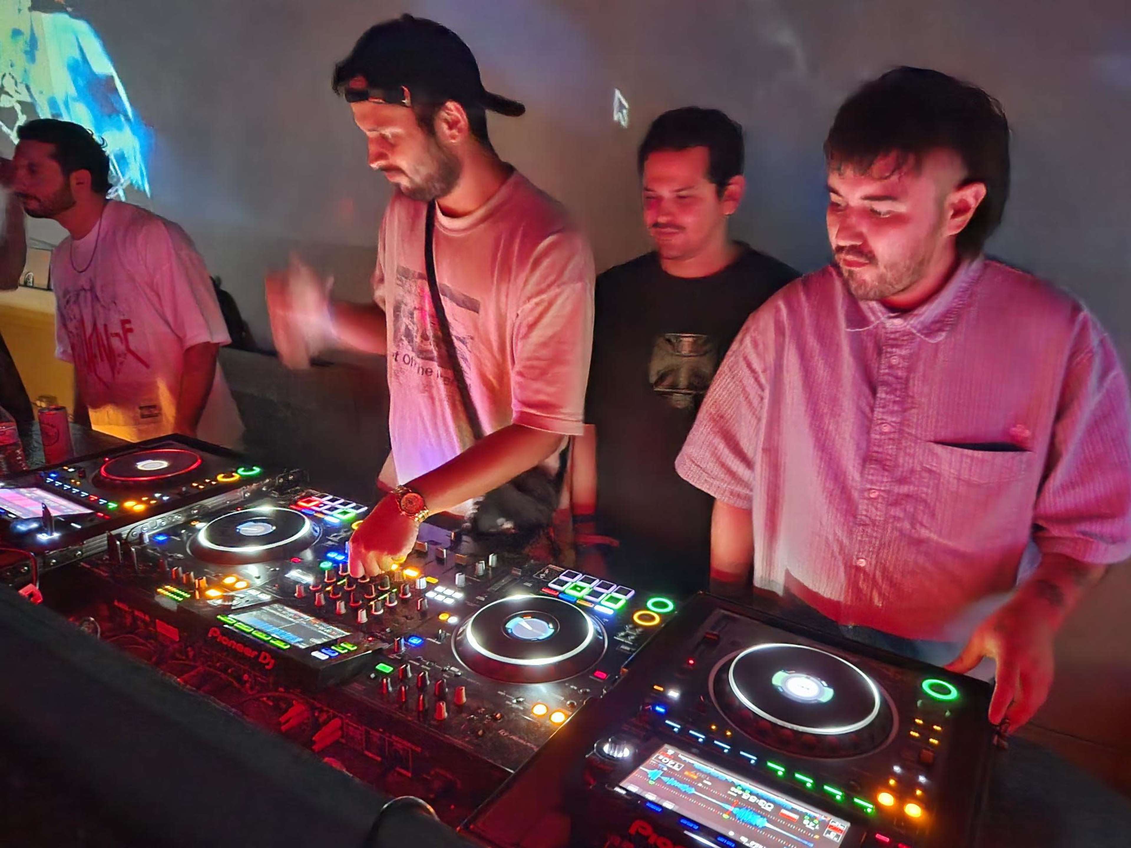 Three individuals are playing music at a DJ booth with colorful lights illuminating the setup.