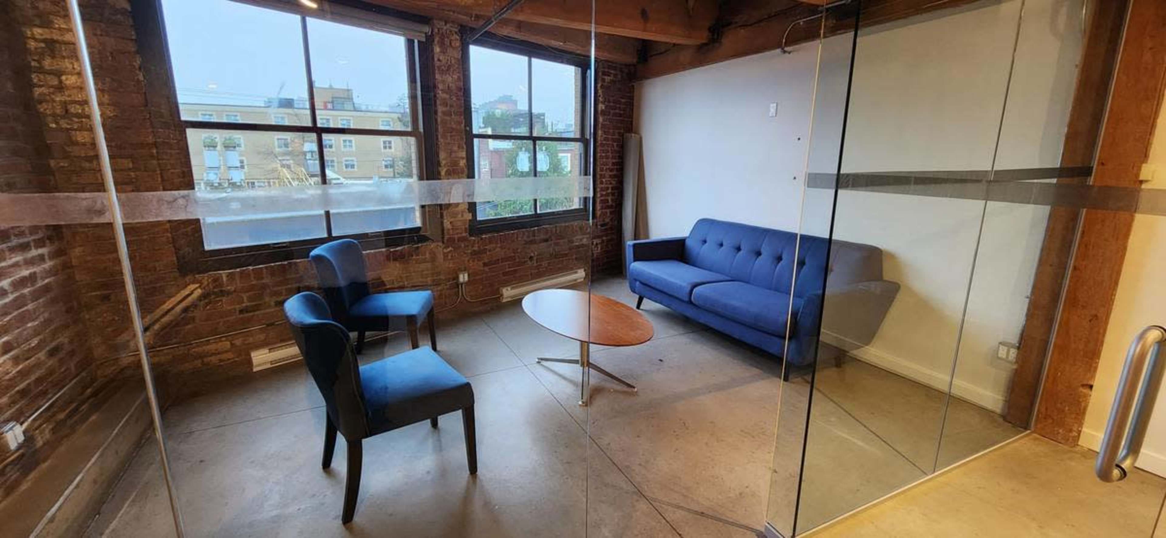 The image shows a small, modern office space with exposed brick walls, featuring a blue sofa, two blue chairs, and a round wooden table.