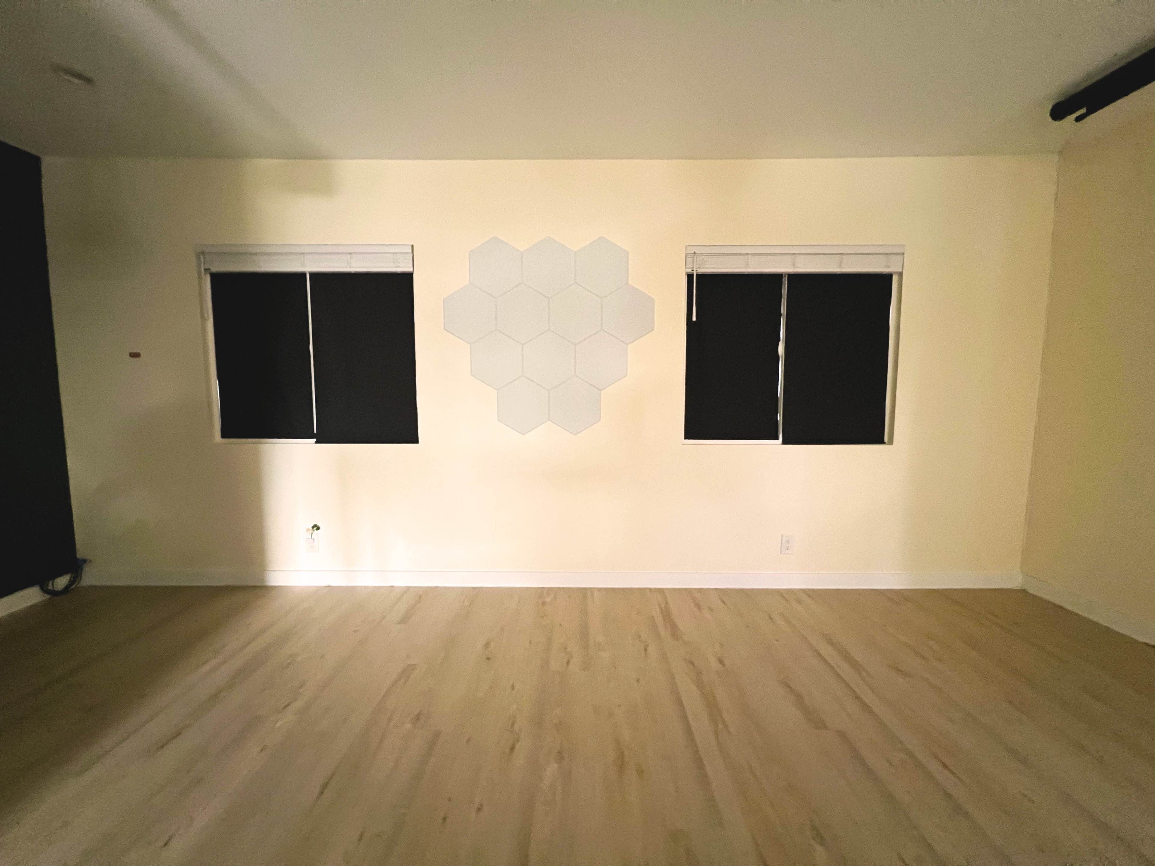 The image shows a room with two windows covered by black blinds, a hexagonal wall feature in the center, and light-colored wooden flooring.