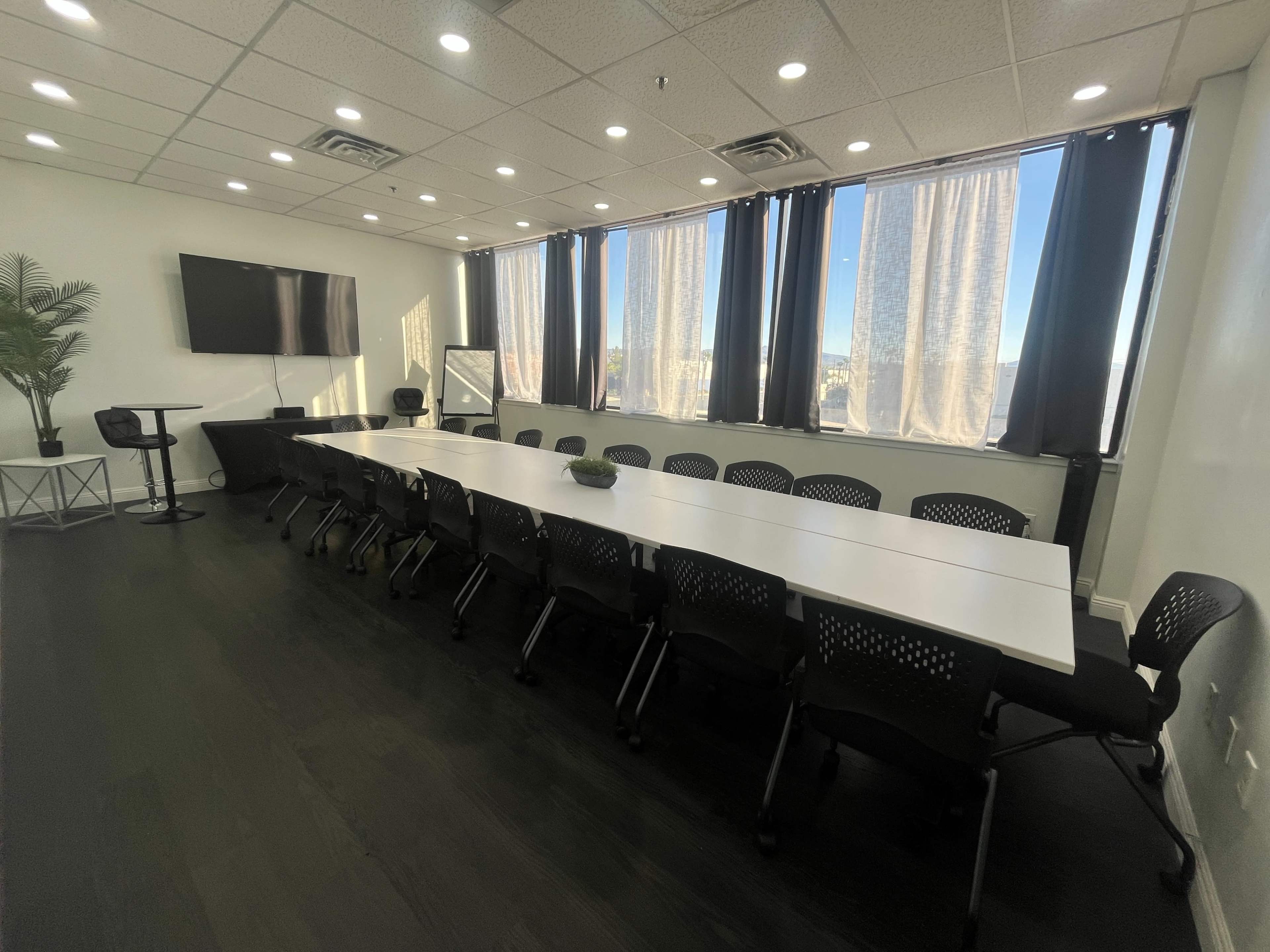 A long conference table with black chairs is arranged in a bright room featuring large windows and a television screen on the wall.