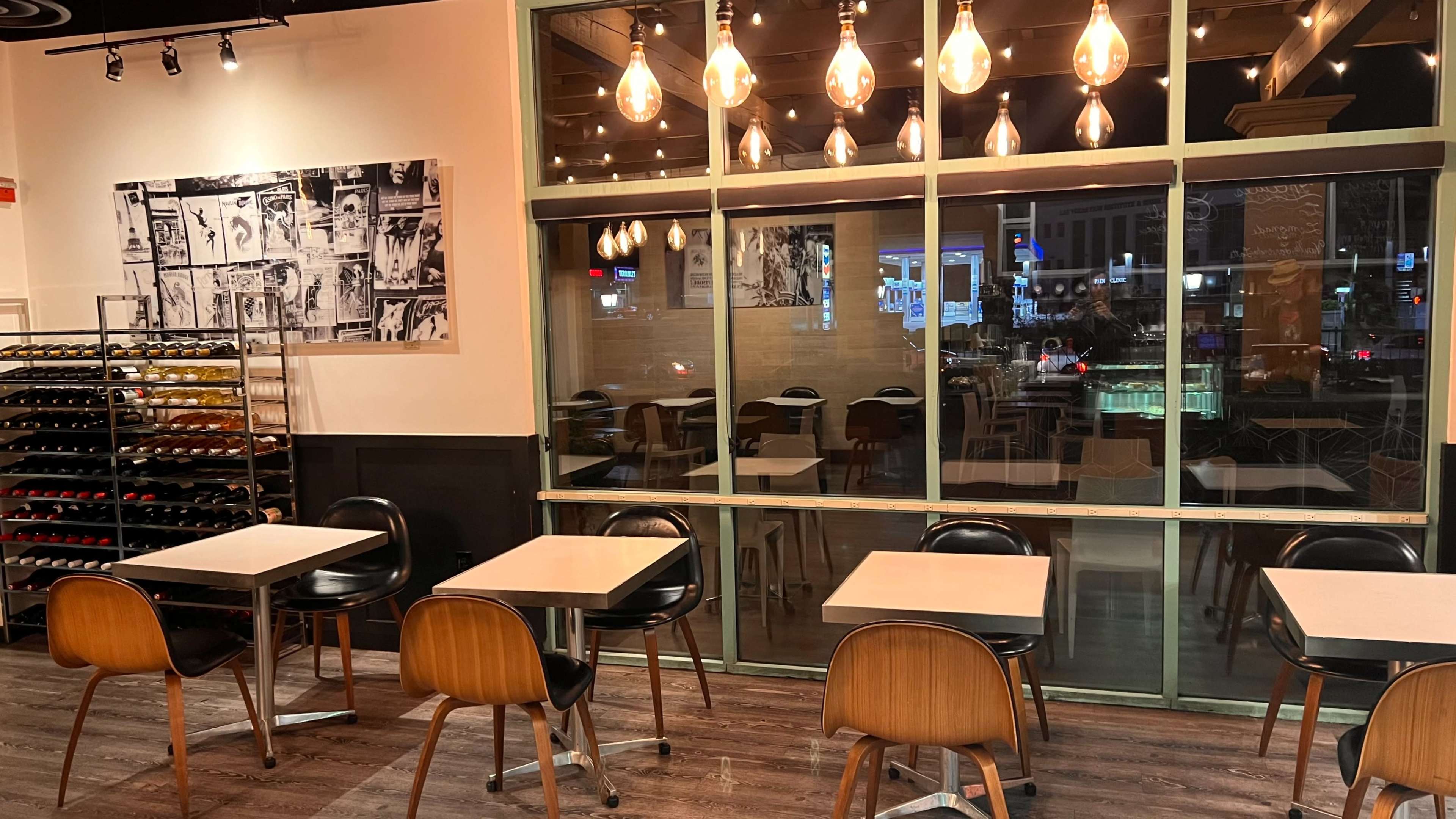 A dining area with wooden chairs, white tables, a wine rack on the side, and large windows displaying an outdoor view illuminated by city lights.