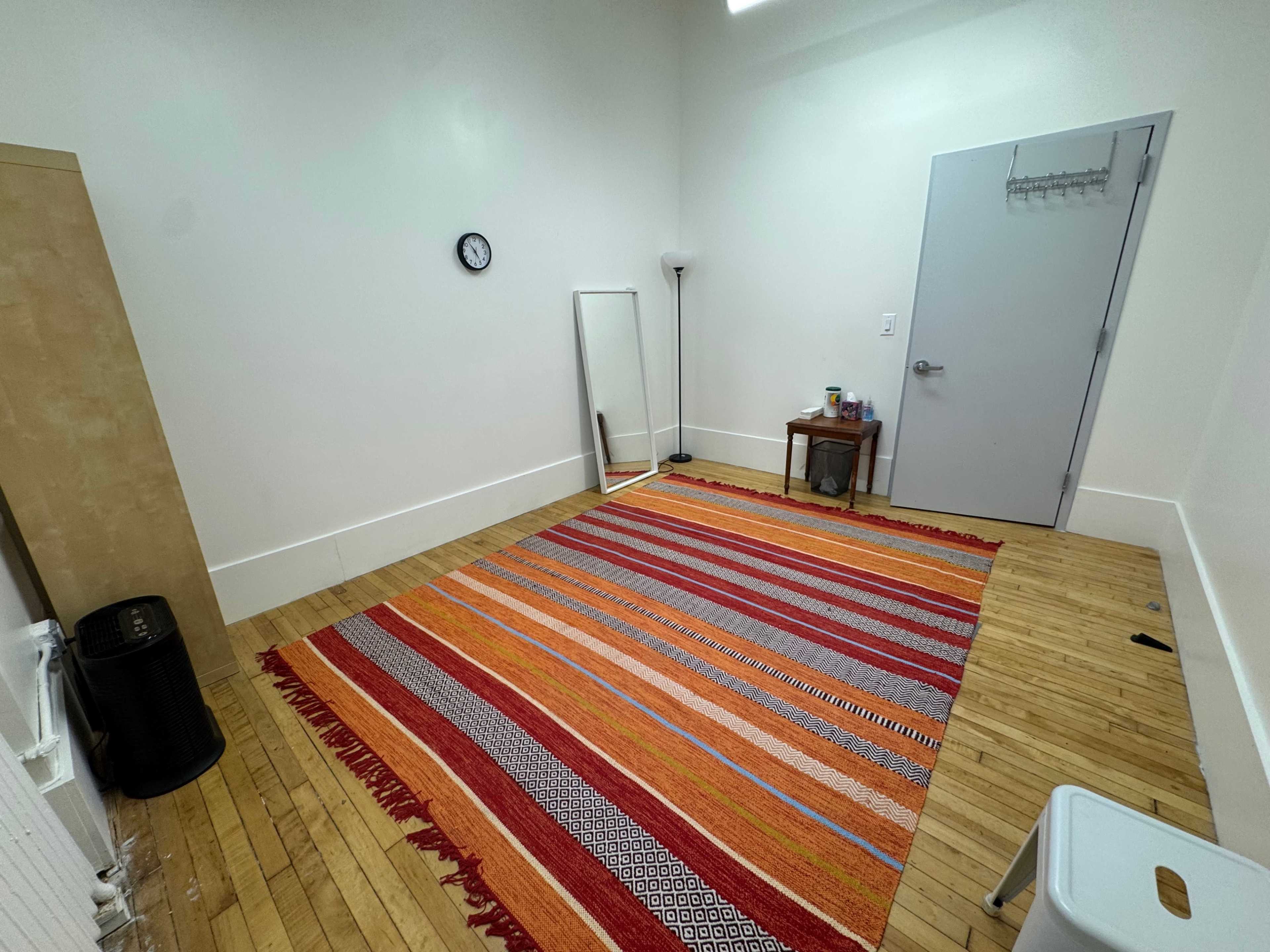 The room features wooden flooring, a large patterned rug, a mirror, a lamp, a clock on the wall, and a simple table with a few items on it.