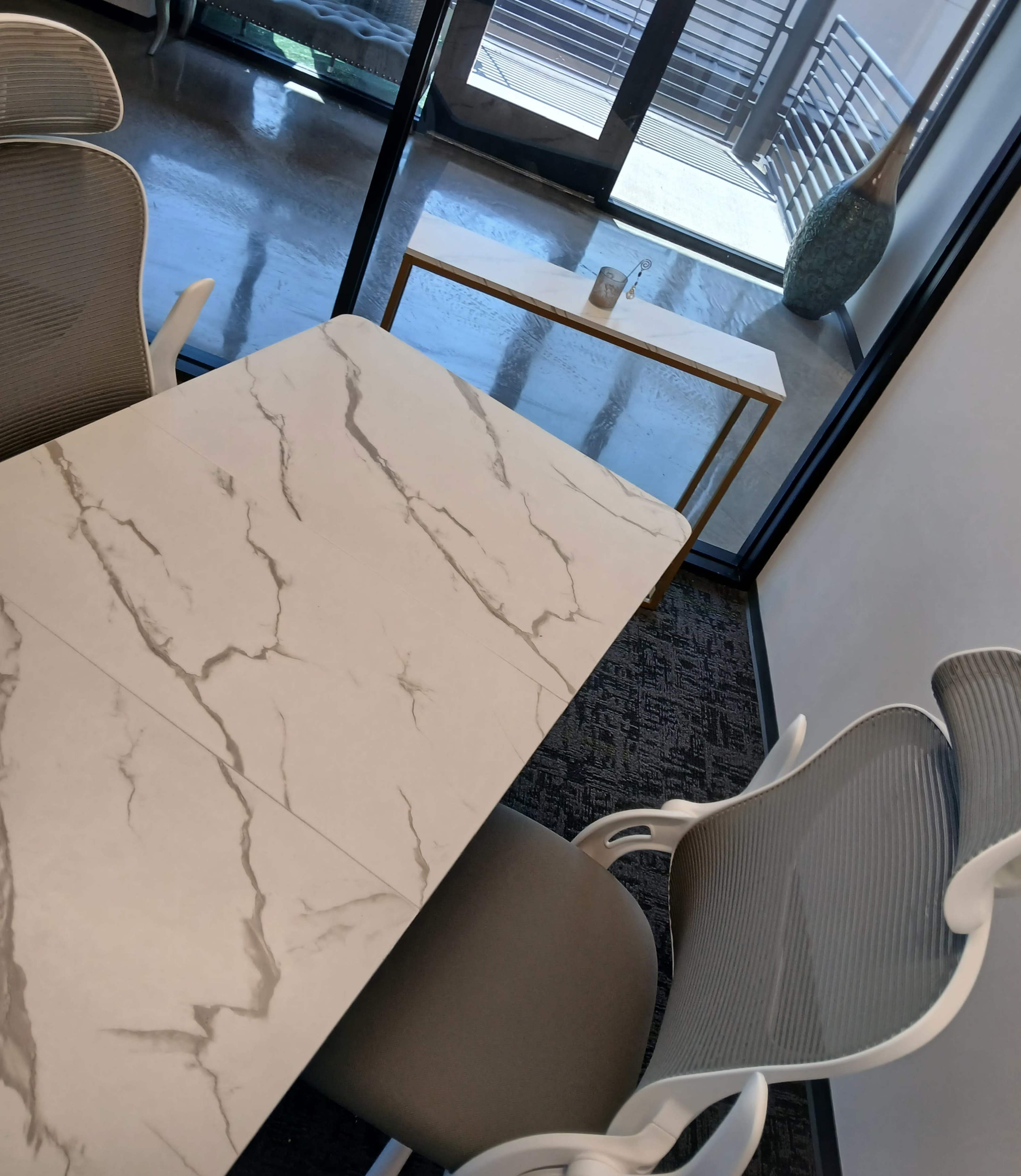 A modern office space features a marble-textured table surrounded by ergonomic chairs, with a decorative table and large windows showing an outdoor area.