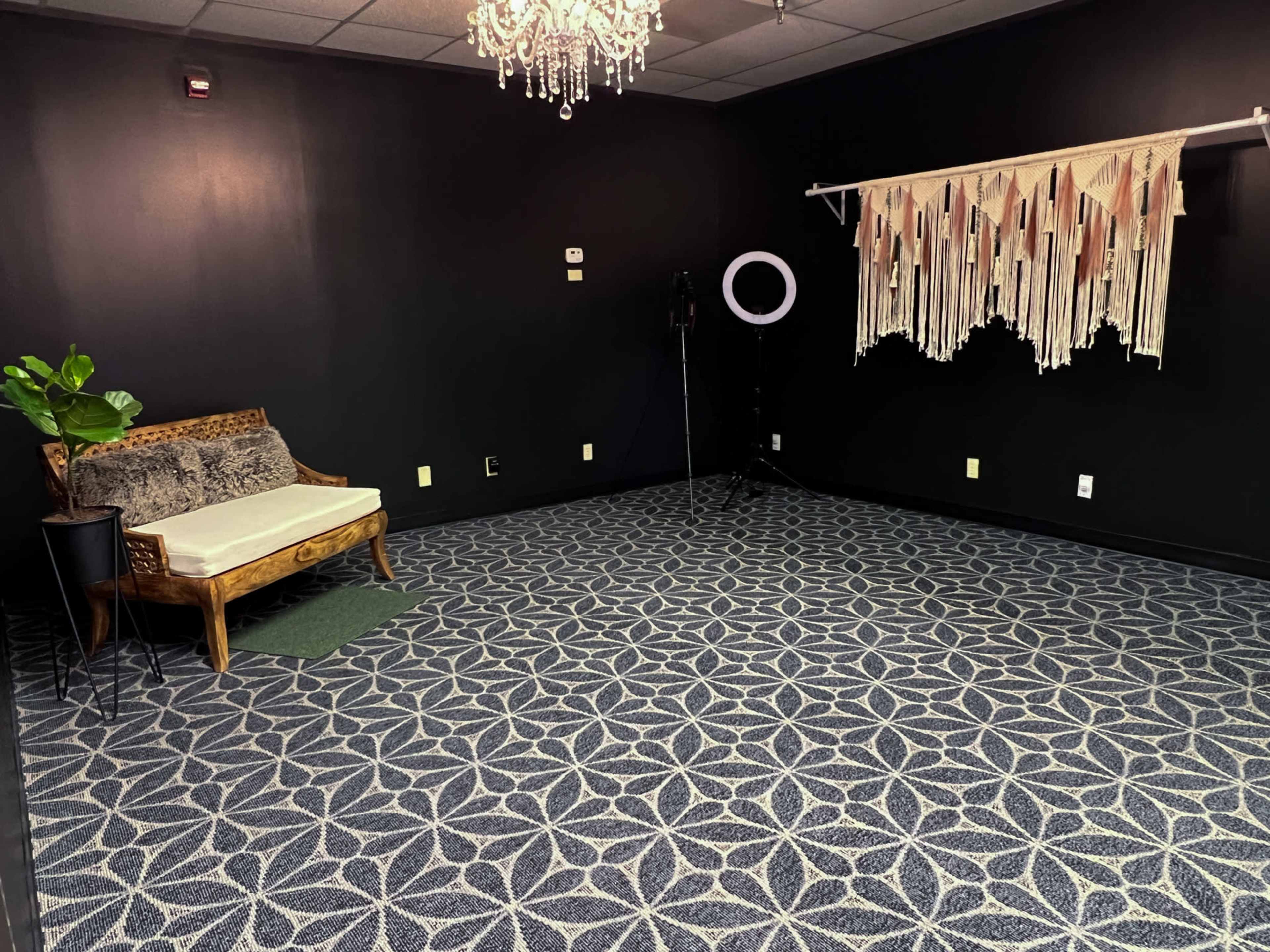 A sparsely furnished room with a patterned carpet, a vintage-style sofa, a plant, and a macramé wall hanging.