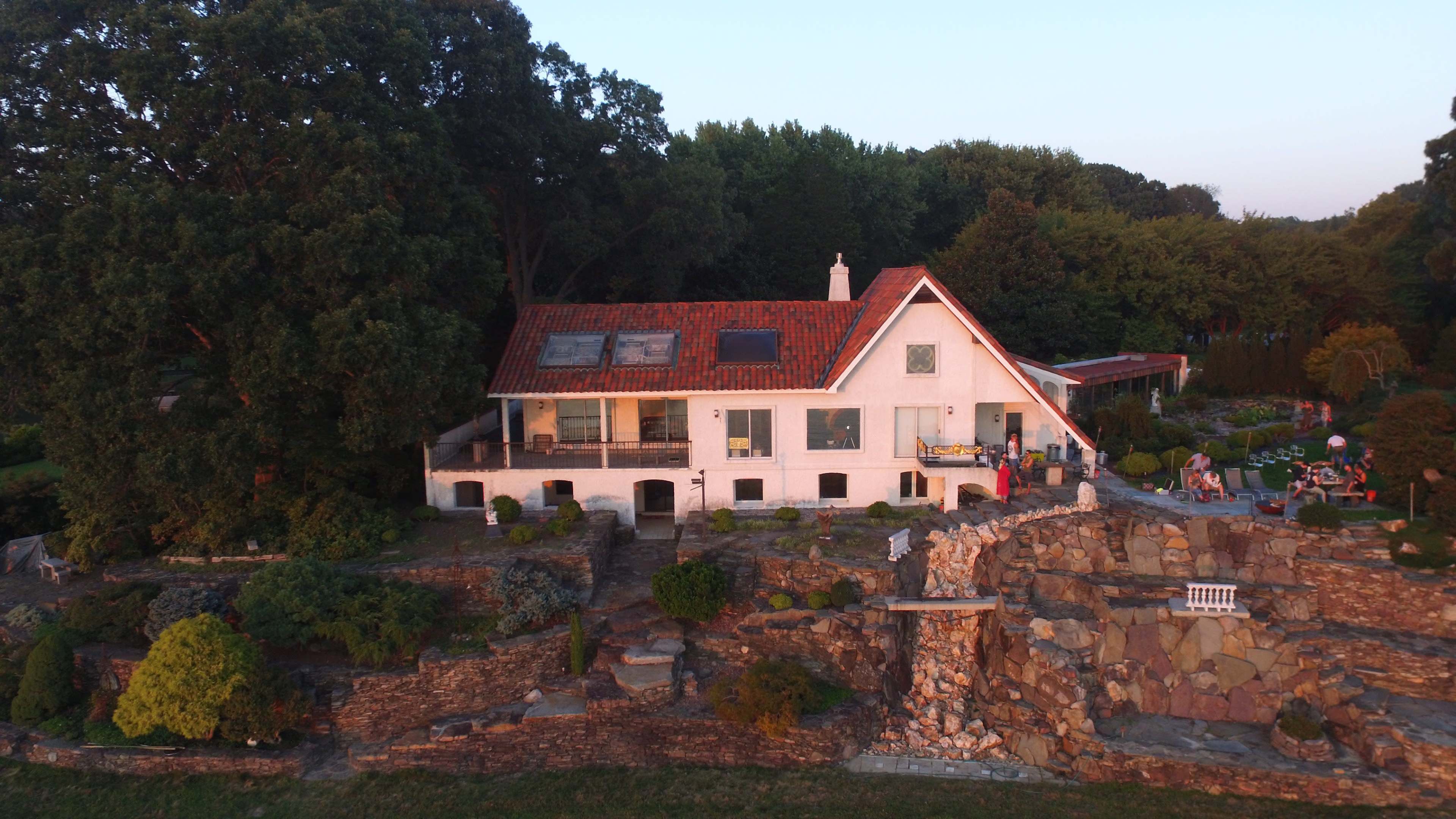 A modern house with a red-tiled roof is situated on a hillside, surrounded by landscaped gardens and trees.