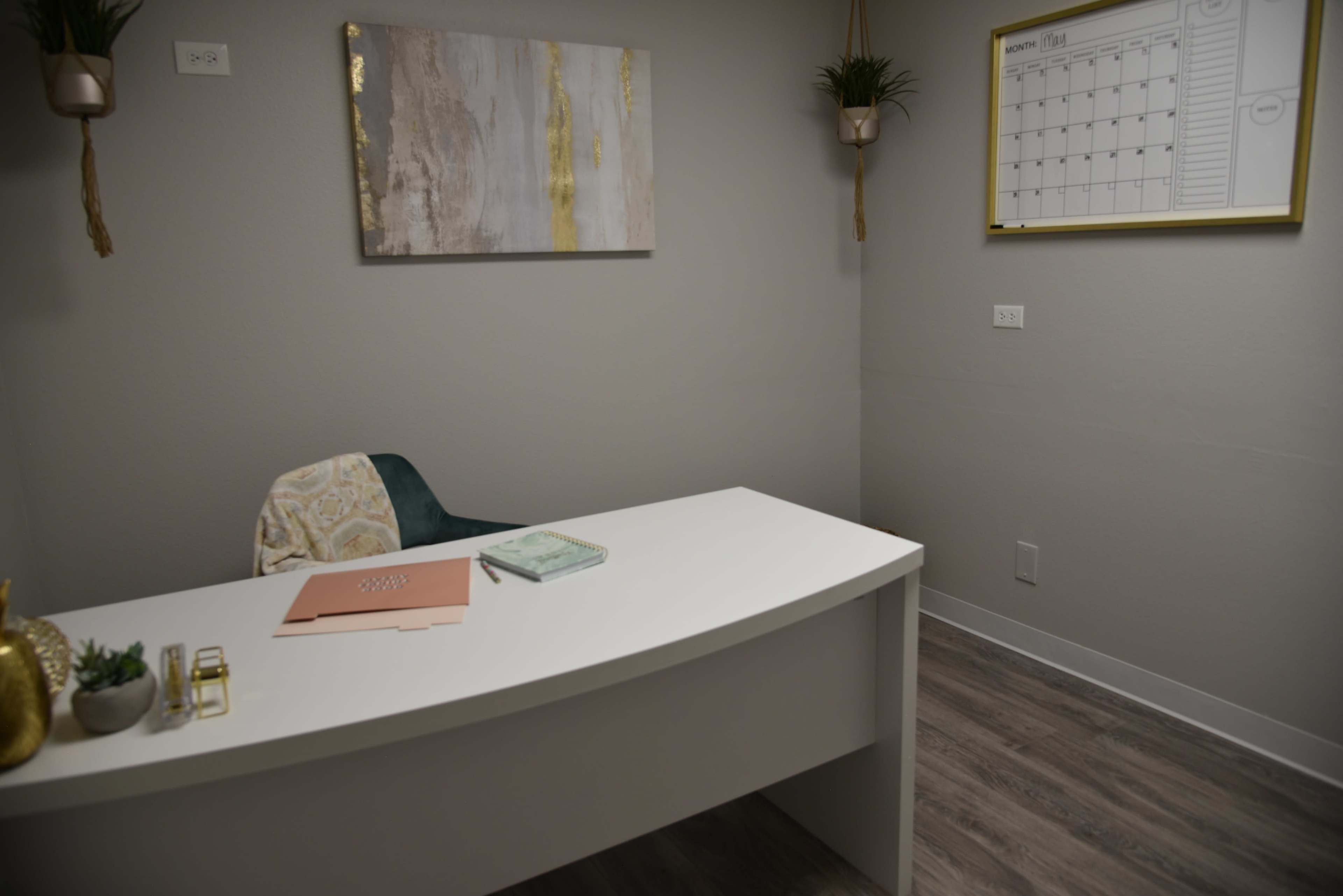 A simple office space features a white desk, a chair with a patterned cushion, a calendar on the wall, and a small plant in a hanging planter.