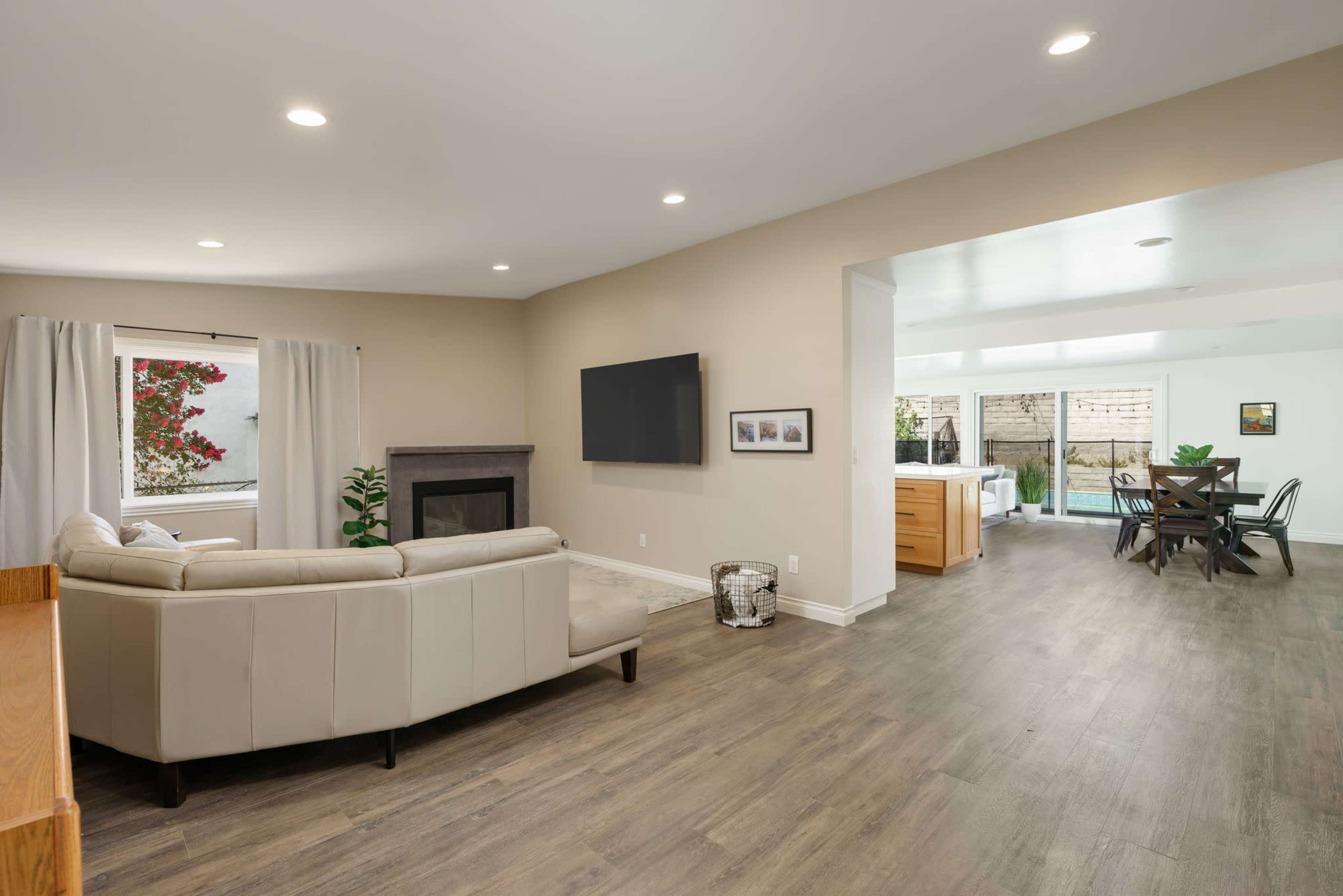 The image shows a modern living room with a beige sectional sofa, a fireplace, and a wall-mounted TV, flowing into a dining area with large windows.