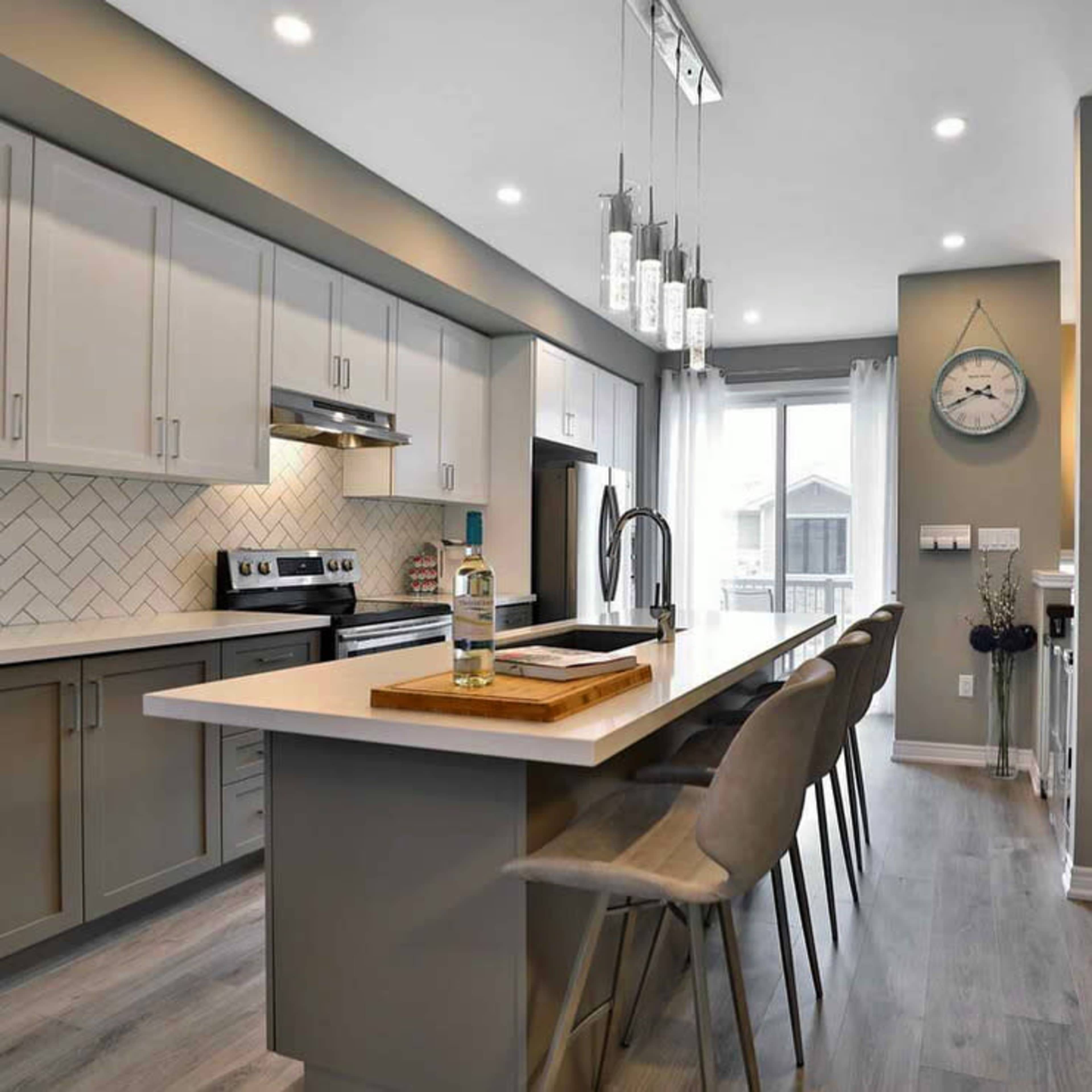 The image shows a modern kitchen featuring light gray cabinetry, a center island with bar seating, and a large window that allows natural light to enter the space.
