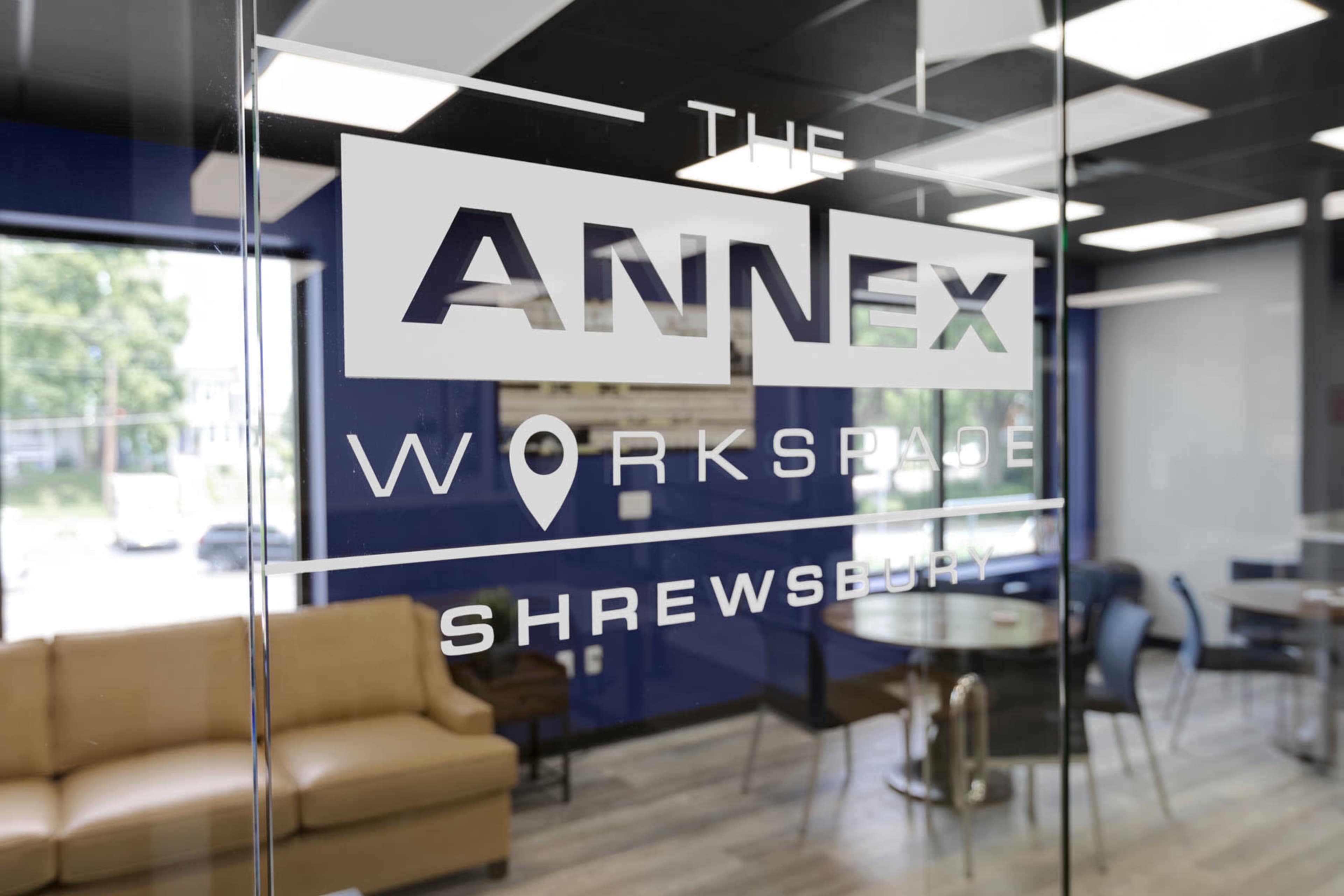A glass door with the words "The Annex Workspace Shrewsbury" etched on it, overlooking a modern office area with a couch and tables.