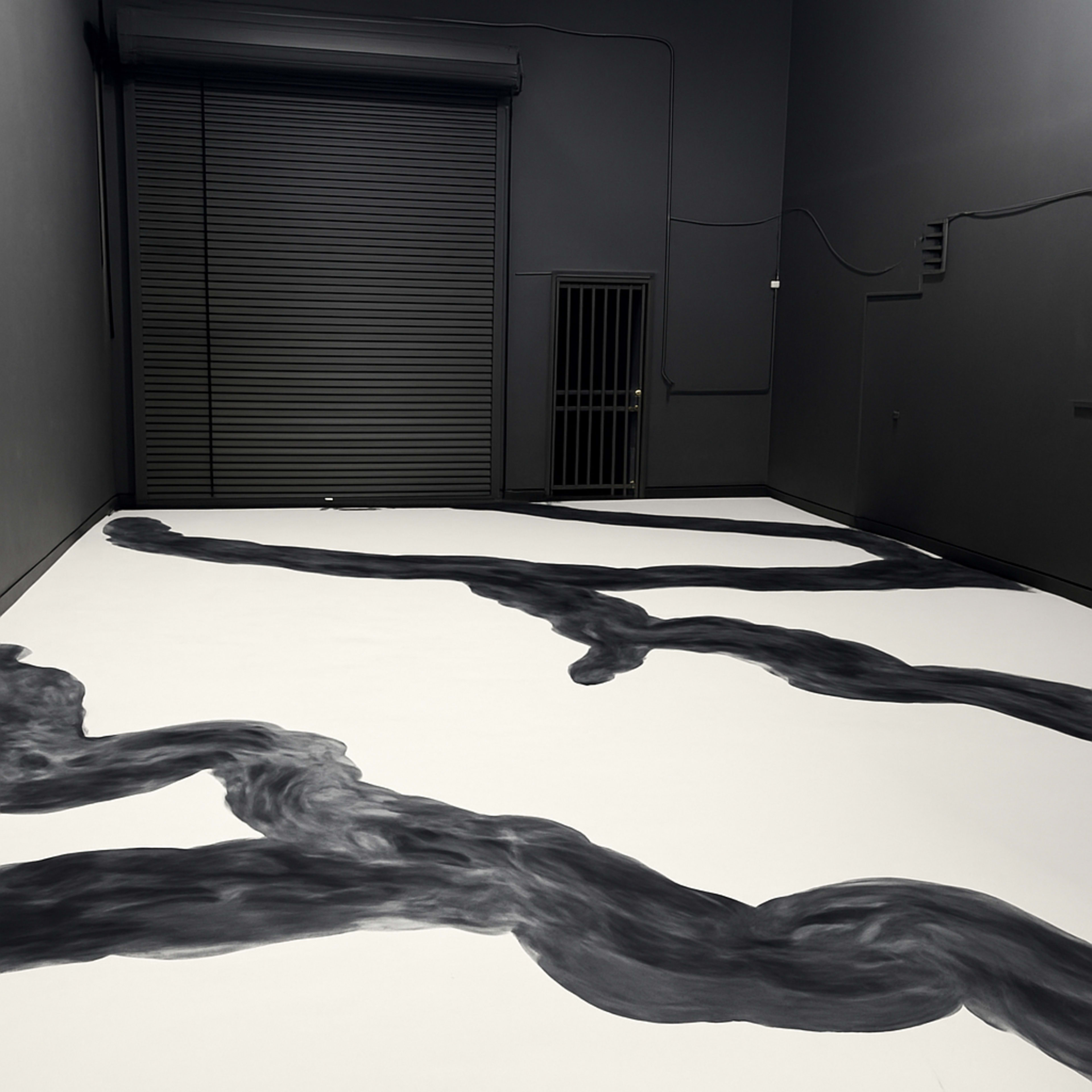 The image shows an empty room with dark, flowing abstract artwork painted on a white floor, contrasting with the grey walls and a metal rolling door at one end.