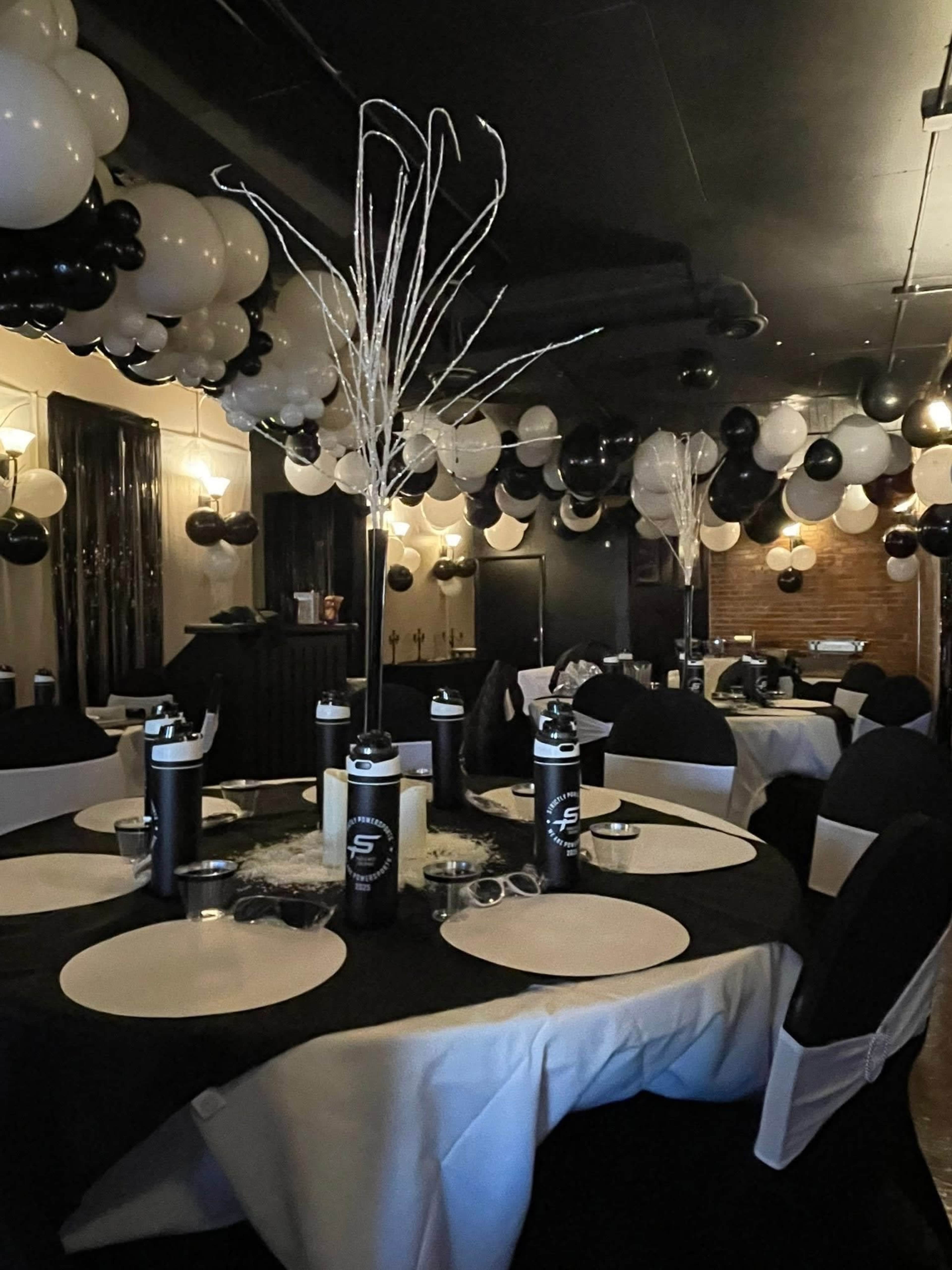 The image shows a banquet hall decorated with black and white balloons, tables set with black and white tablecloths, and centerpieces featuring silver branches.