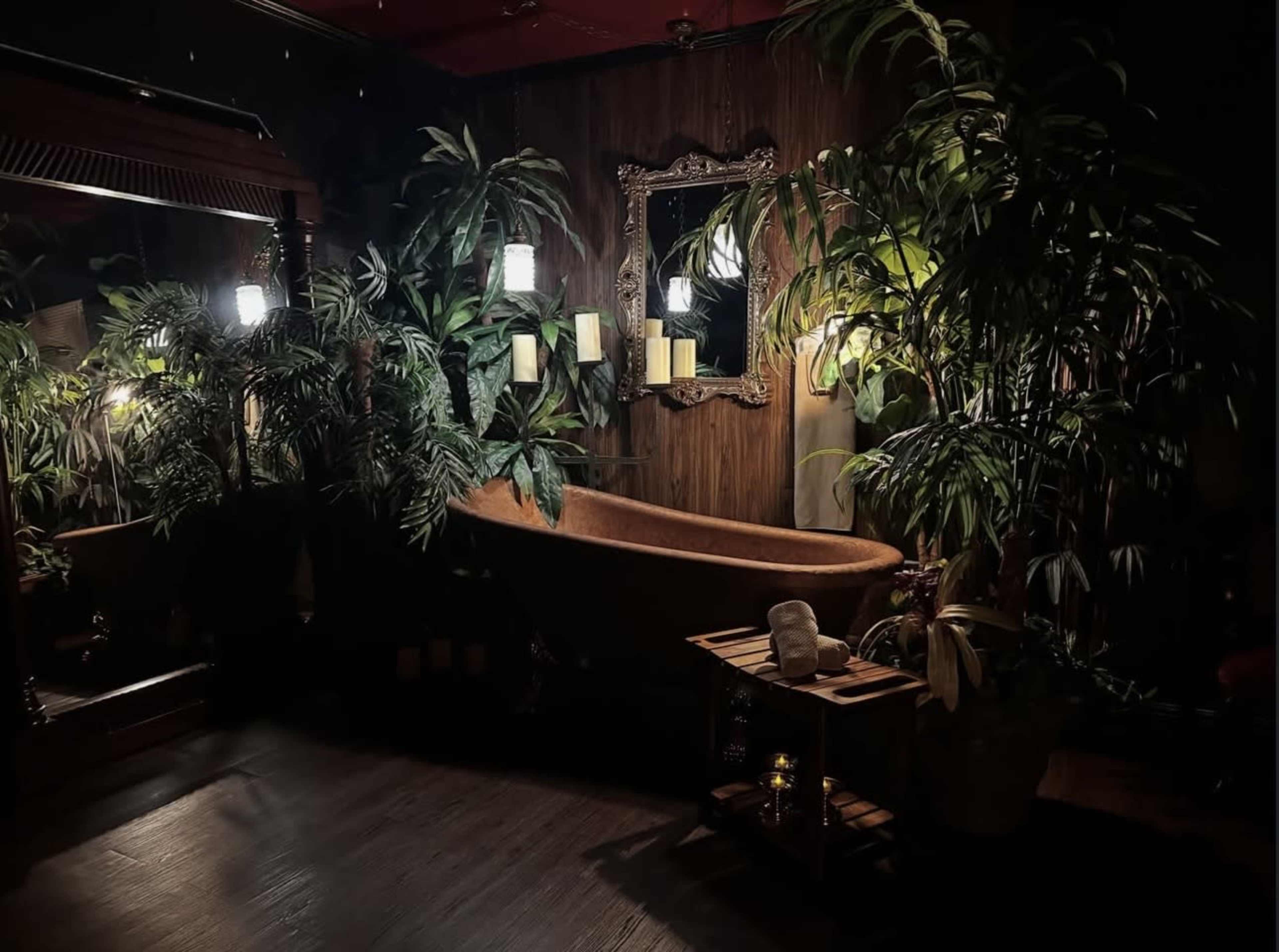 The image shows an intimate bathroom setting featuring a large, freestanding bathtub surrounded by various lush green plants and soft lighting.