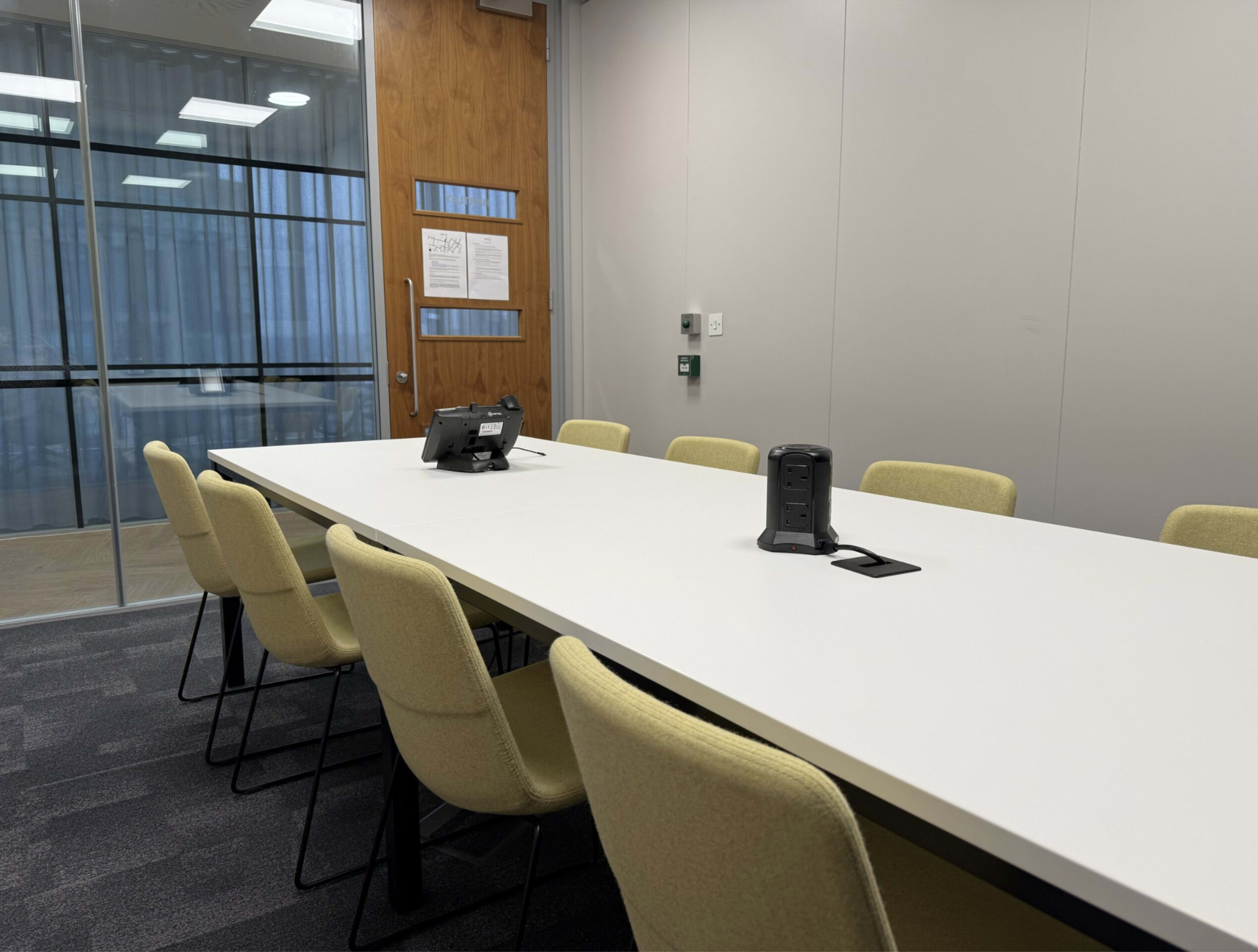 A long, white conference table with several yellow chairs surrounds it, positioned in a modern meeting room with glass walls.