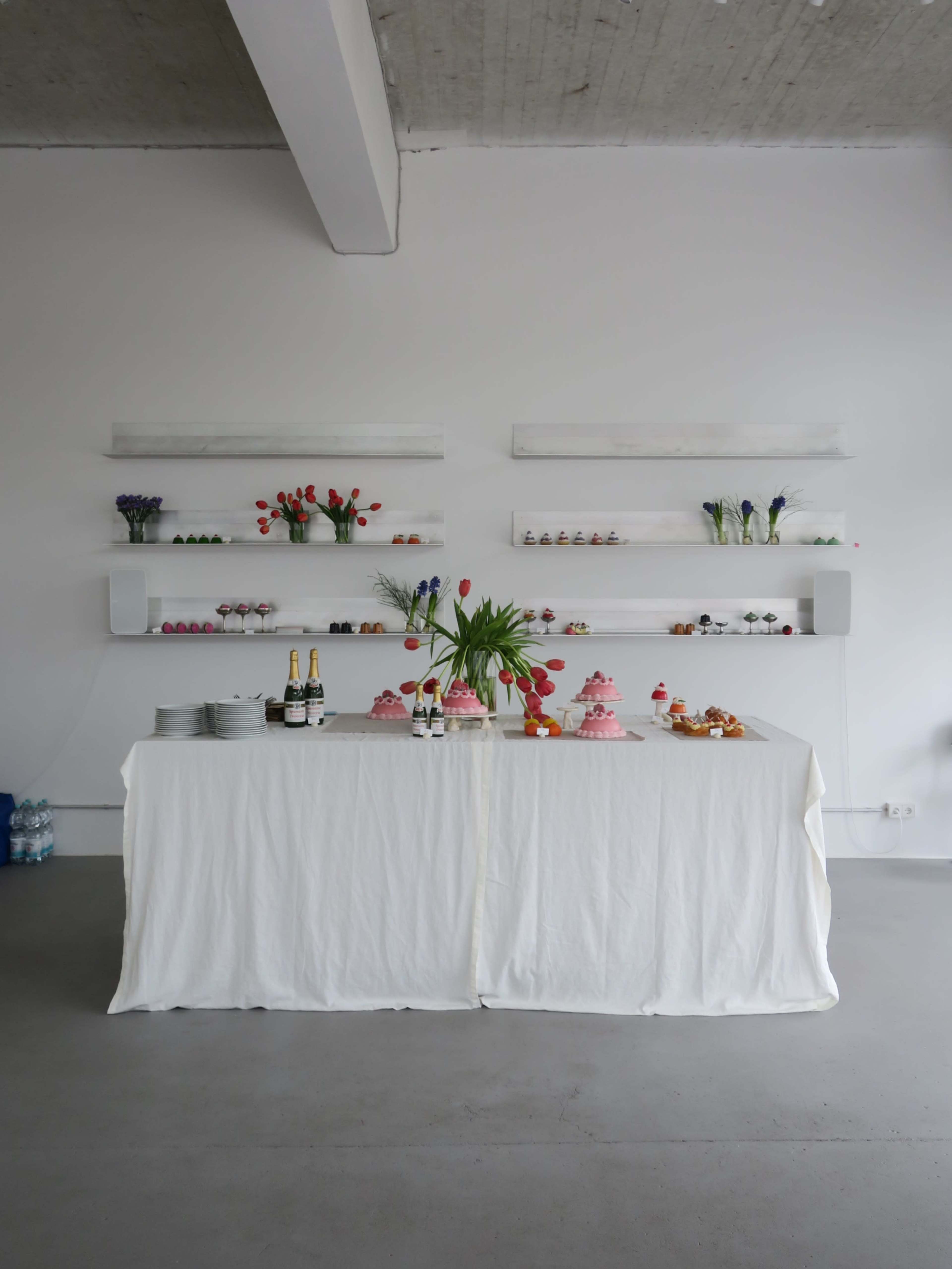 A white table is set with a tablecloth, displaying various desserts and decorative flowers against a minimalist backdrop.