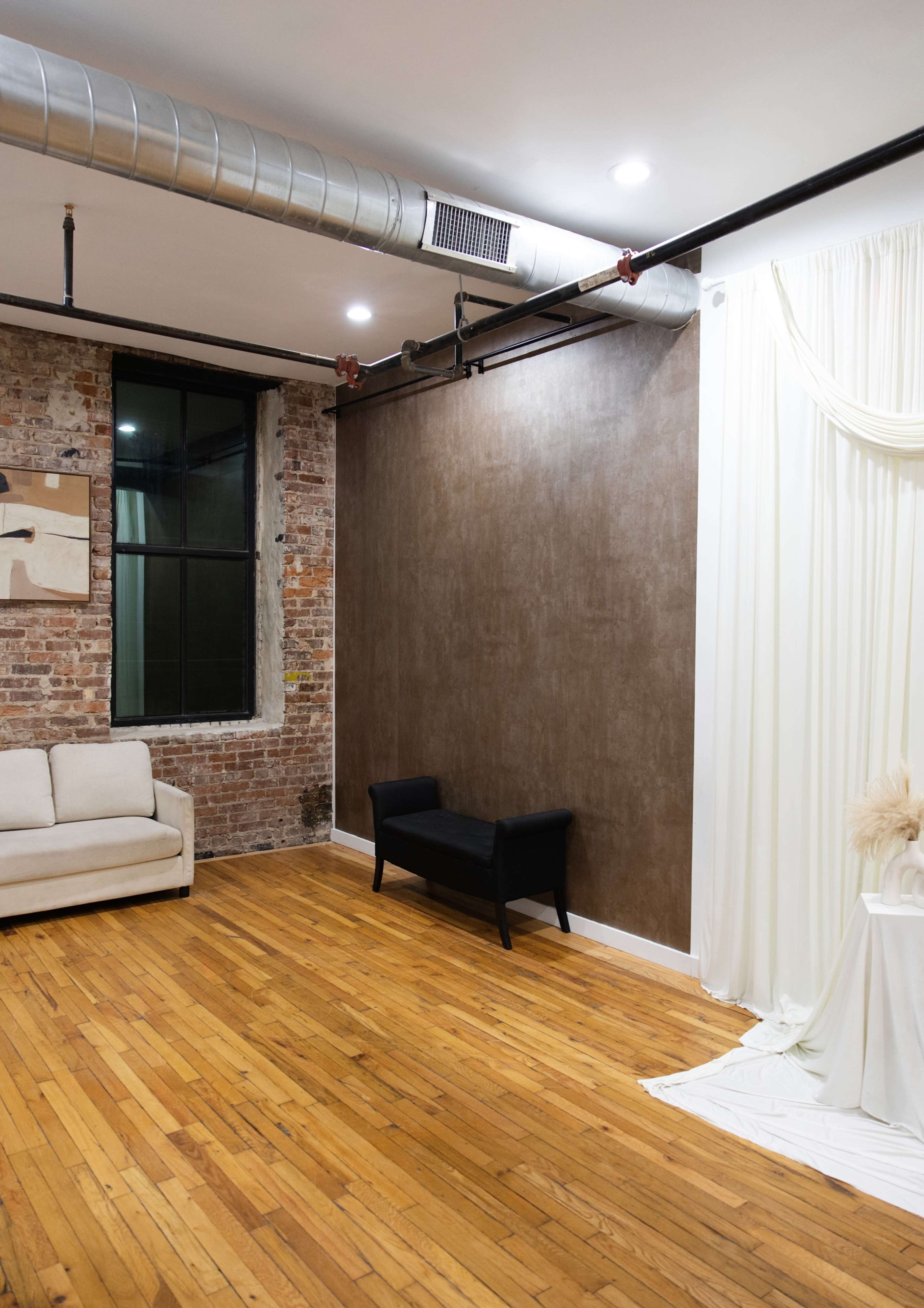 The image shows a spacious interior featuring a wooden floor, a brick wall, a black couch, and a light-colored drapery against a brown wall.