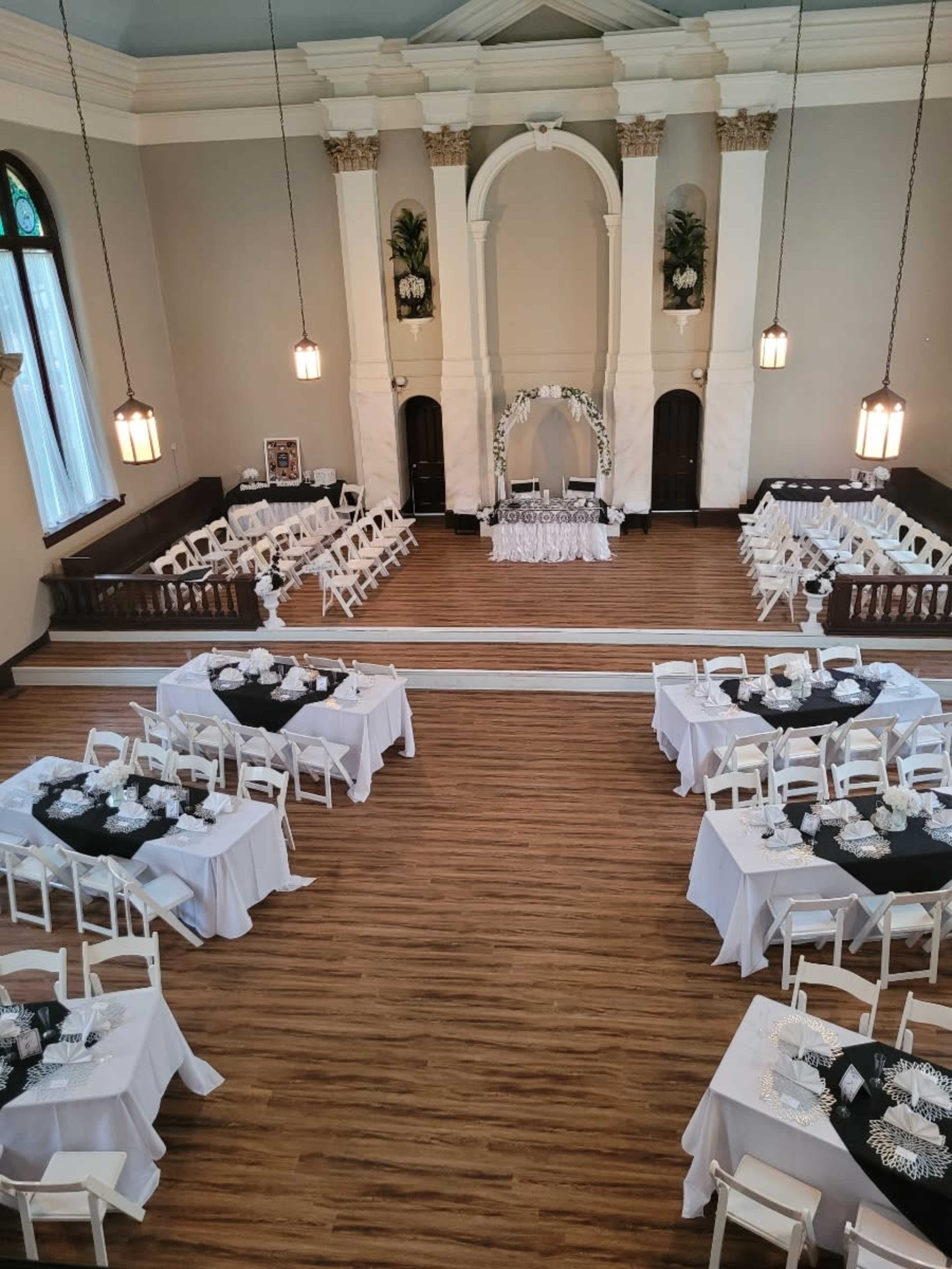 The image shows a large banquet hall set up for an event, with several white tables arranged in two groups facing a central stage adorned with floral decor.