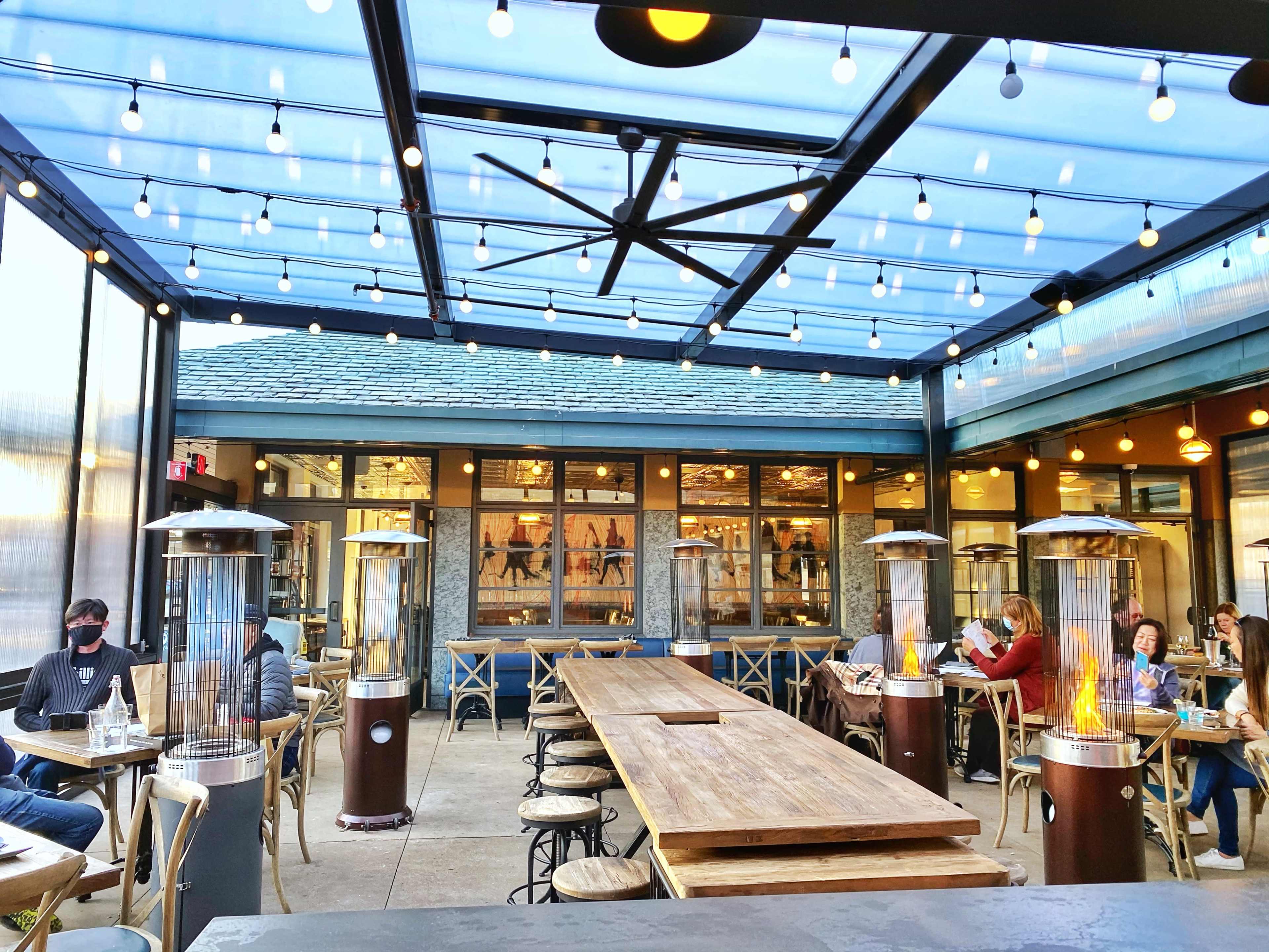 A spacious outdoor dining area with modern heating lamps, string lights, and a large wooden table surrounded by patrons.