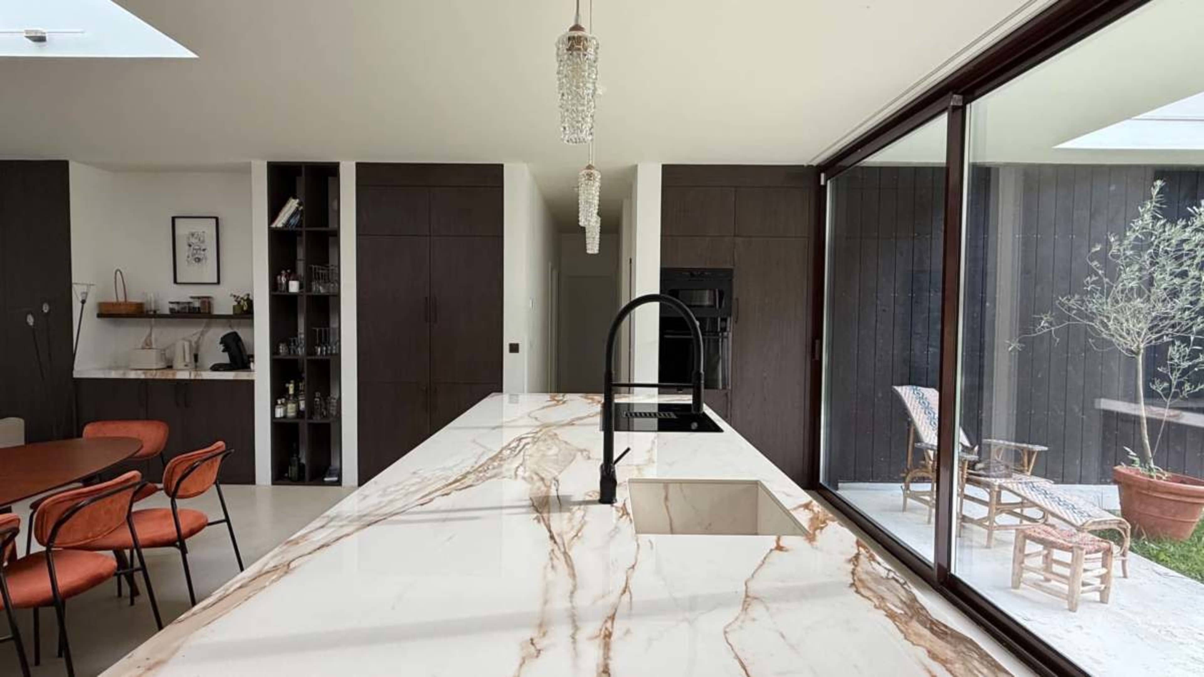 The image shows a modern kitchen with a marble countertop, a sleek black faucet, and large windows that open to a small outdoor area with plants.