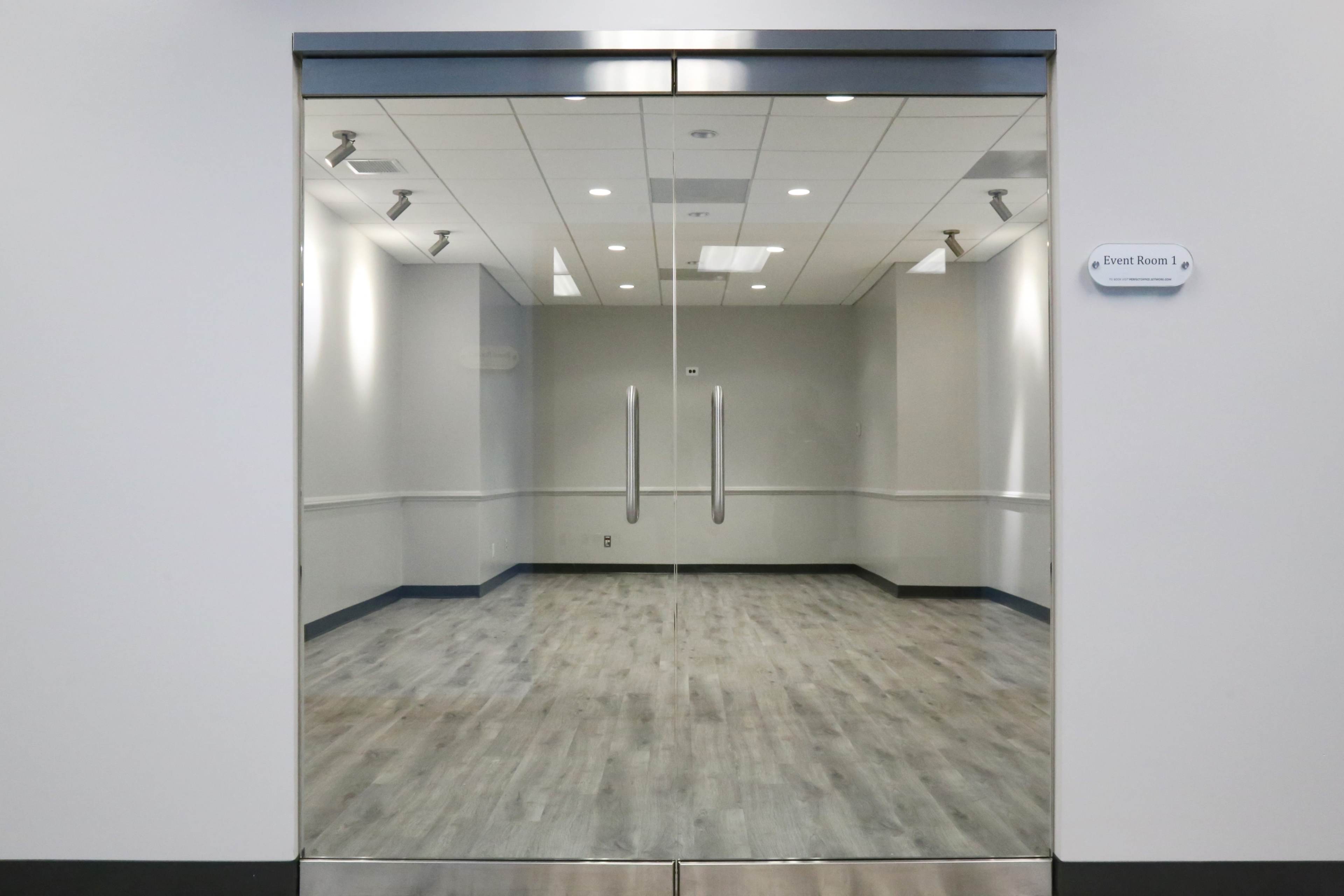 The image shows a glass entrance to an empty event room with grey walls and a wooden floor.