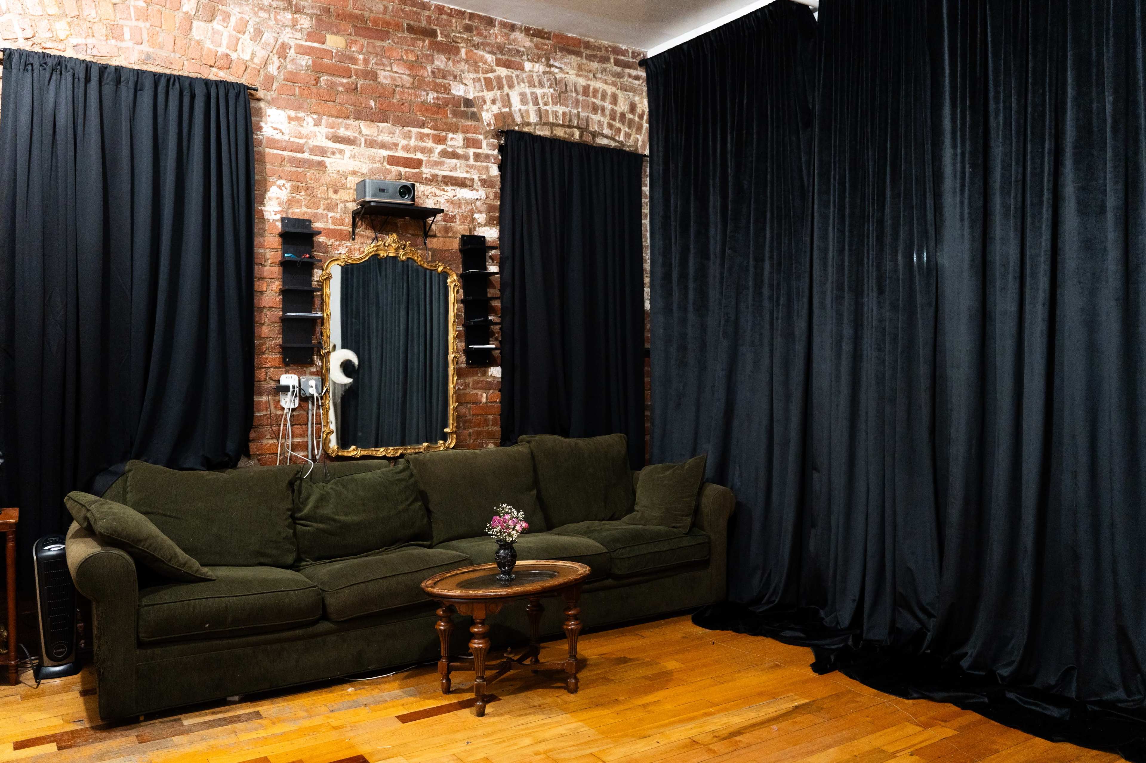 The image shows a cozy living room with a dark green sofa, a round wooden table, a vintage mirror, and thick black curtains covering the windows against a backdrop of exposed brick walls.