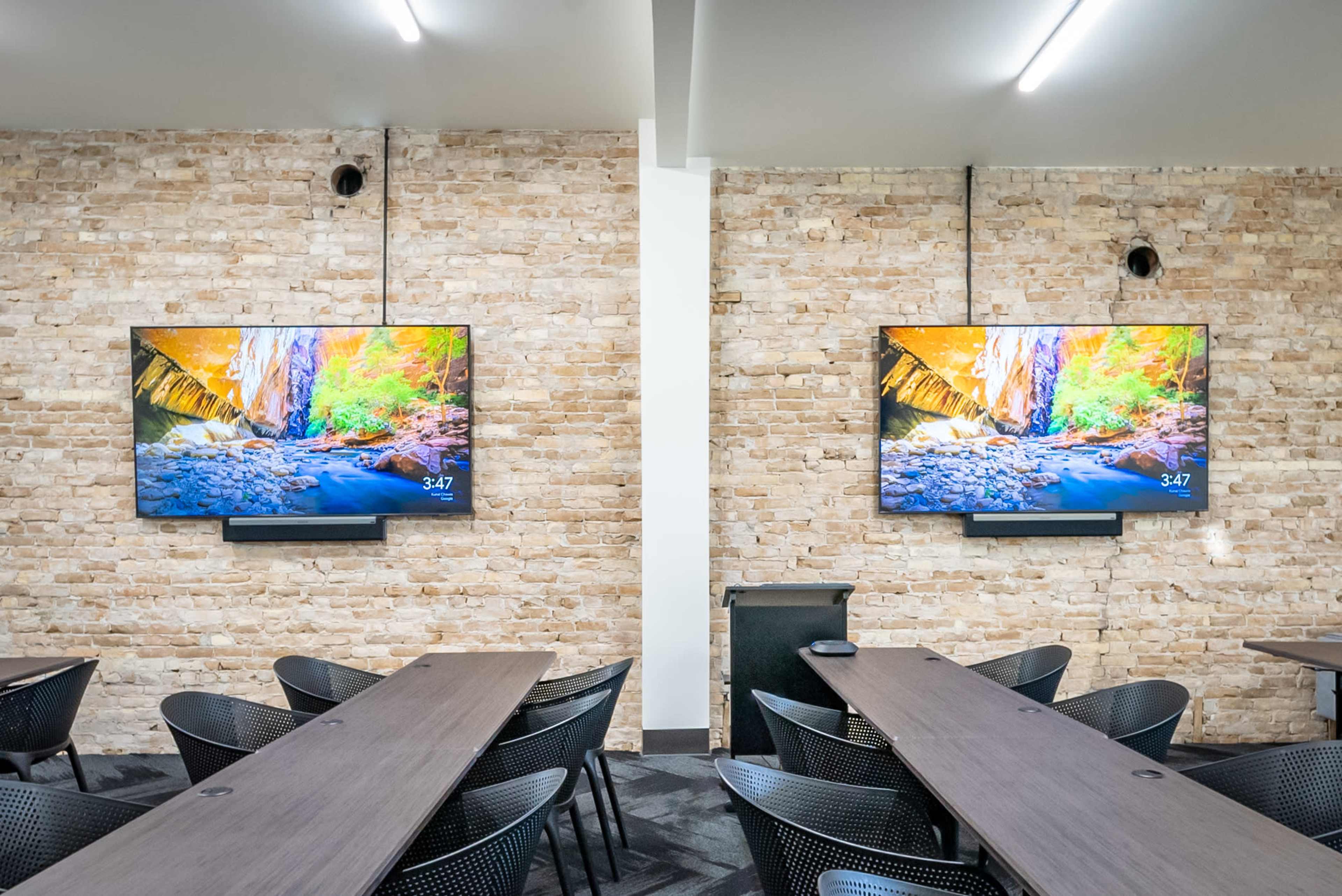 A conference room with two wall-mounted televisions displaying a nature scene, flanked by long tables and black chairs, against a light brick wall.