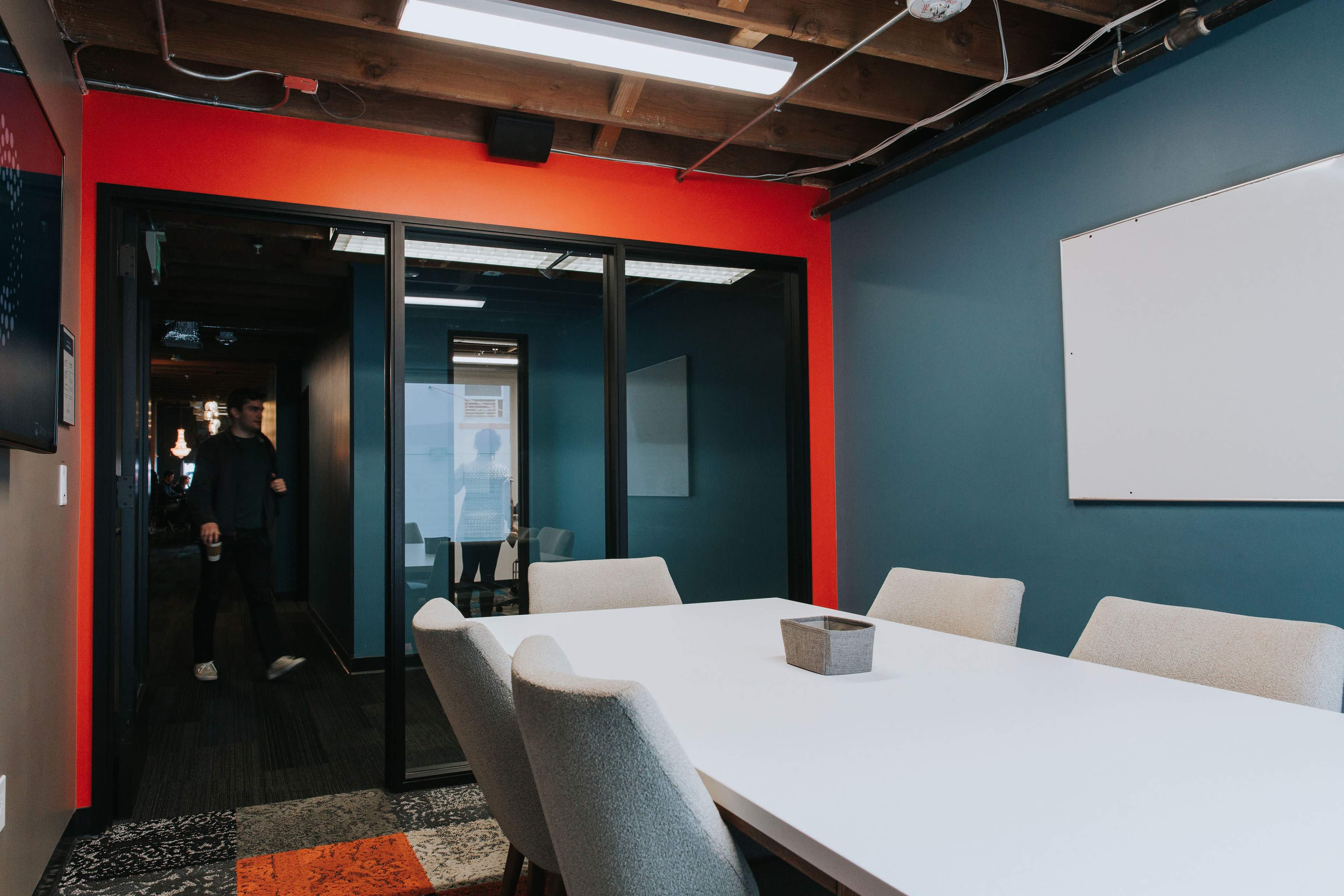 A conference room features a long white table surrounded by light-colored chairs, with a glass wall leading to another room visible in the background.