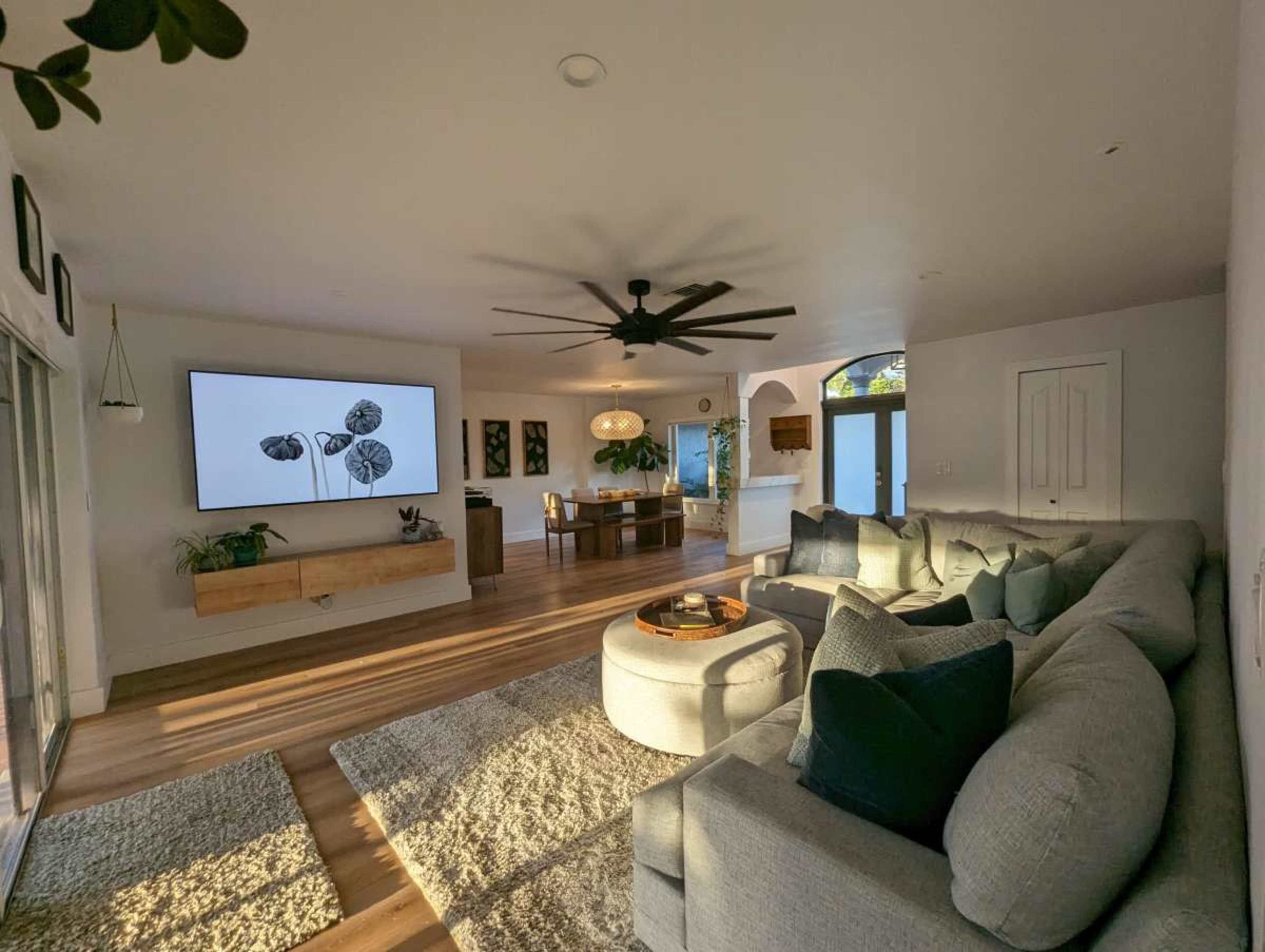 A modern living room features a large gray sectional sofa, a circular coffee table, and a flat-screen TV mounted on the wall, with an adjacent dining area visible in the background.