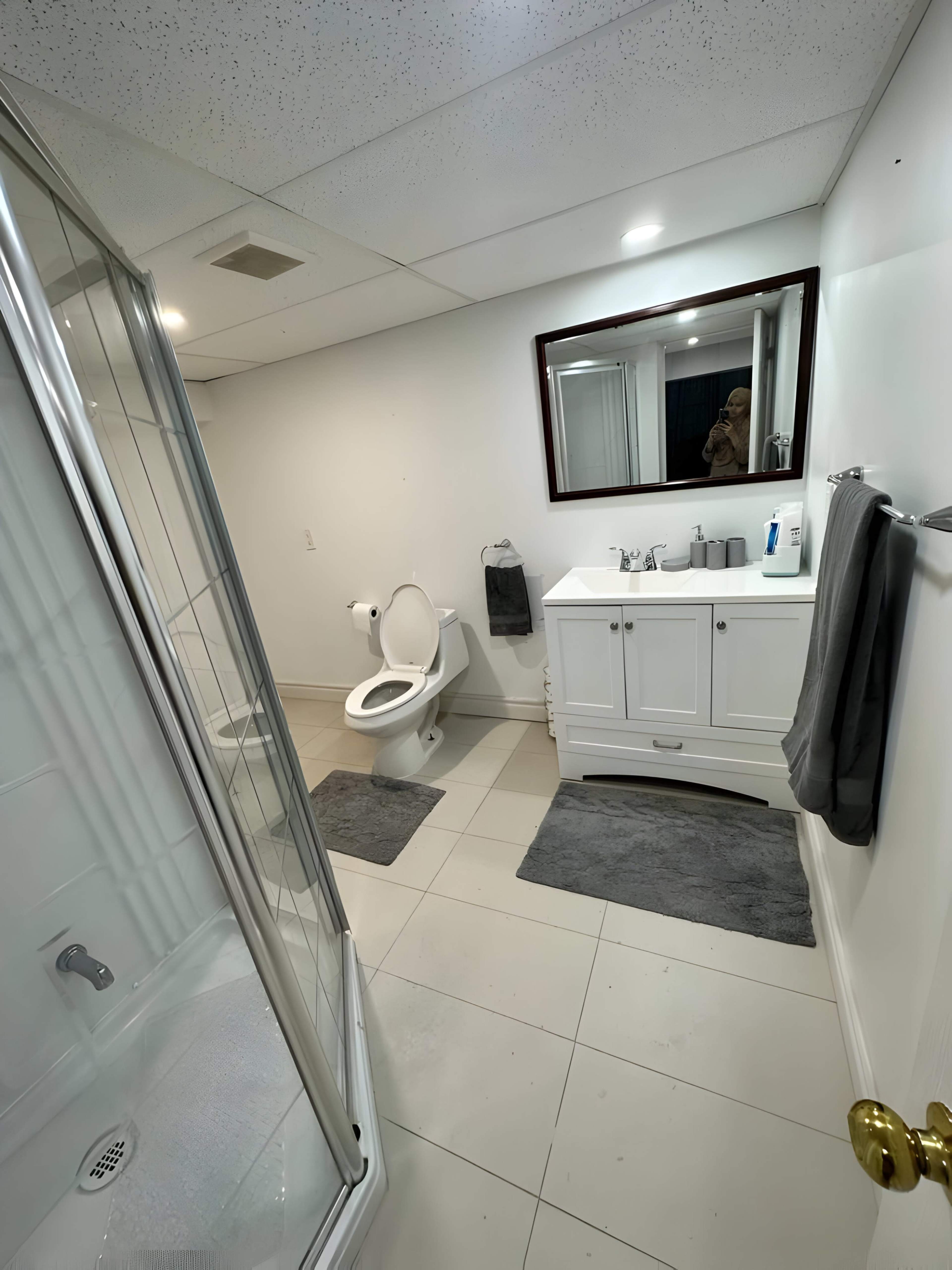 A bathroom with a shower, toilet, sink vanity, and a large mirror, all set against neutral-colored walls and tiled flooring.