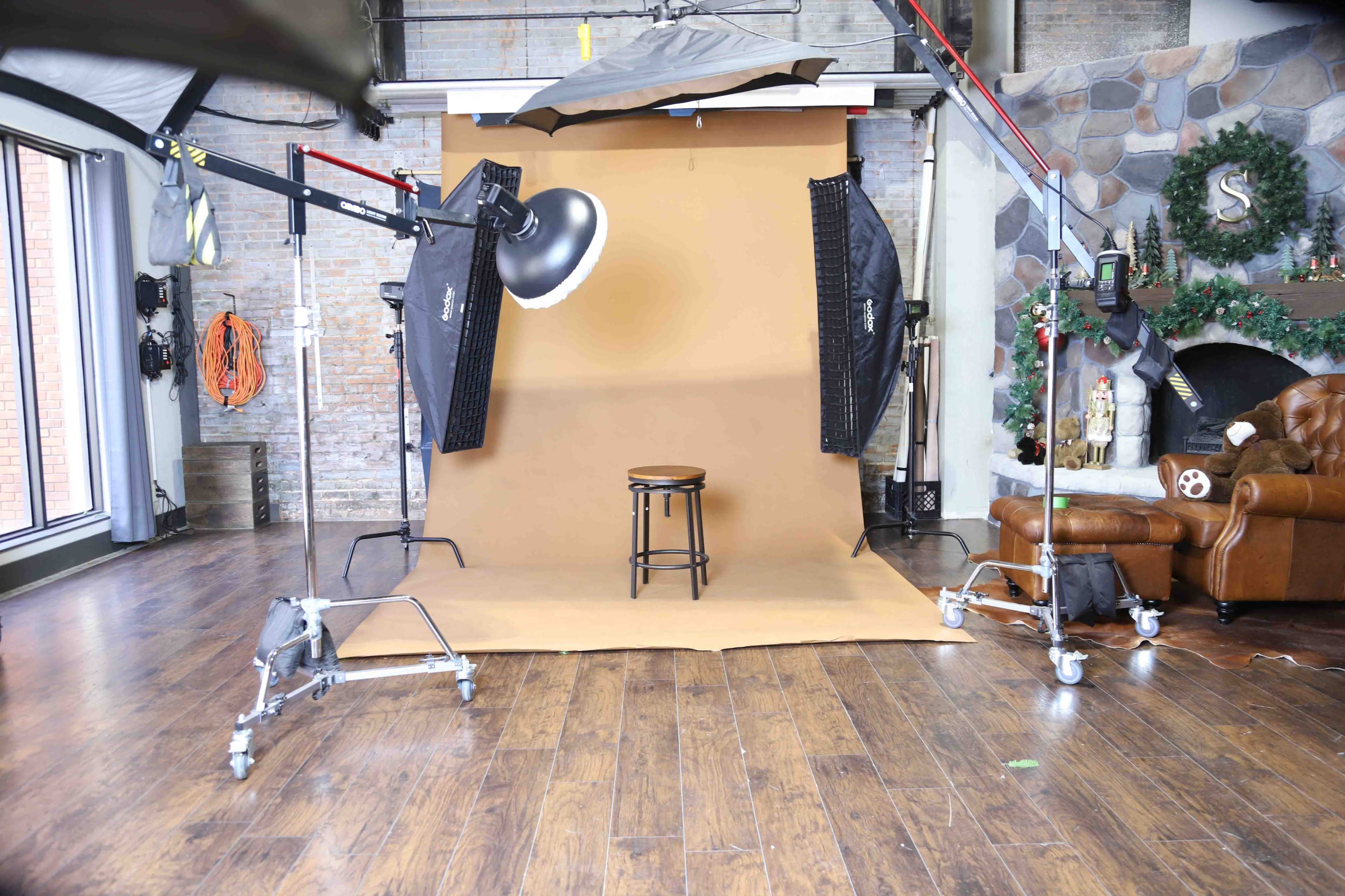 A photography studio setup featuring a brown backdrop, a stool in the center, and various lighting equipment arranged around the scene.