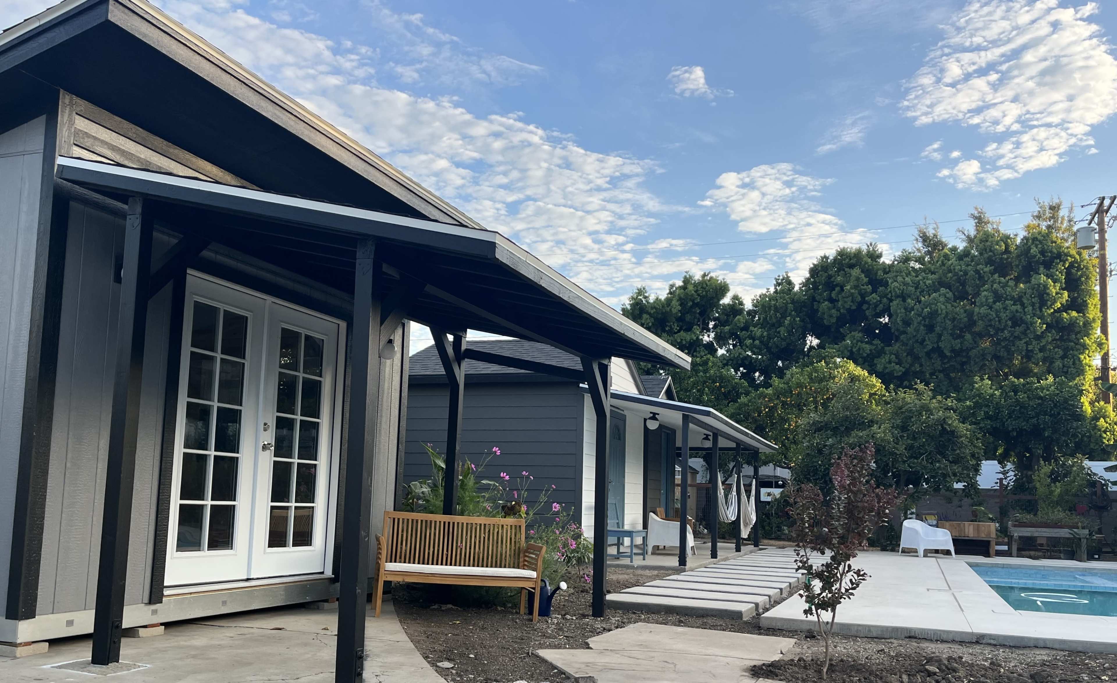 The image shows a residential backyard with a modern house, a wooden bench, a stone pathway, and a swimming pool surrounded by greenery under a partly cloudy sky.