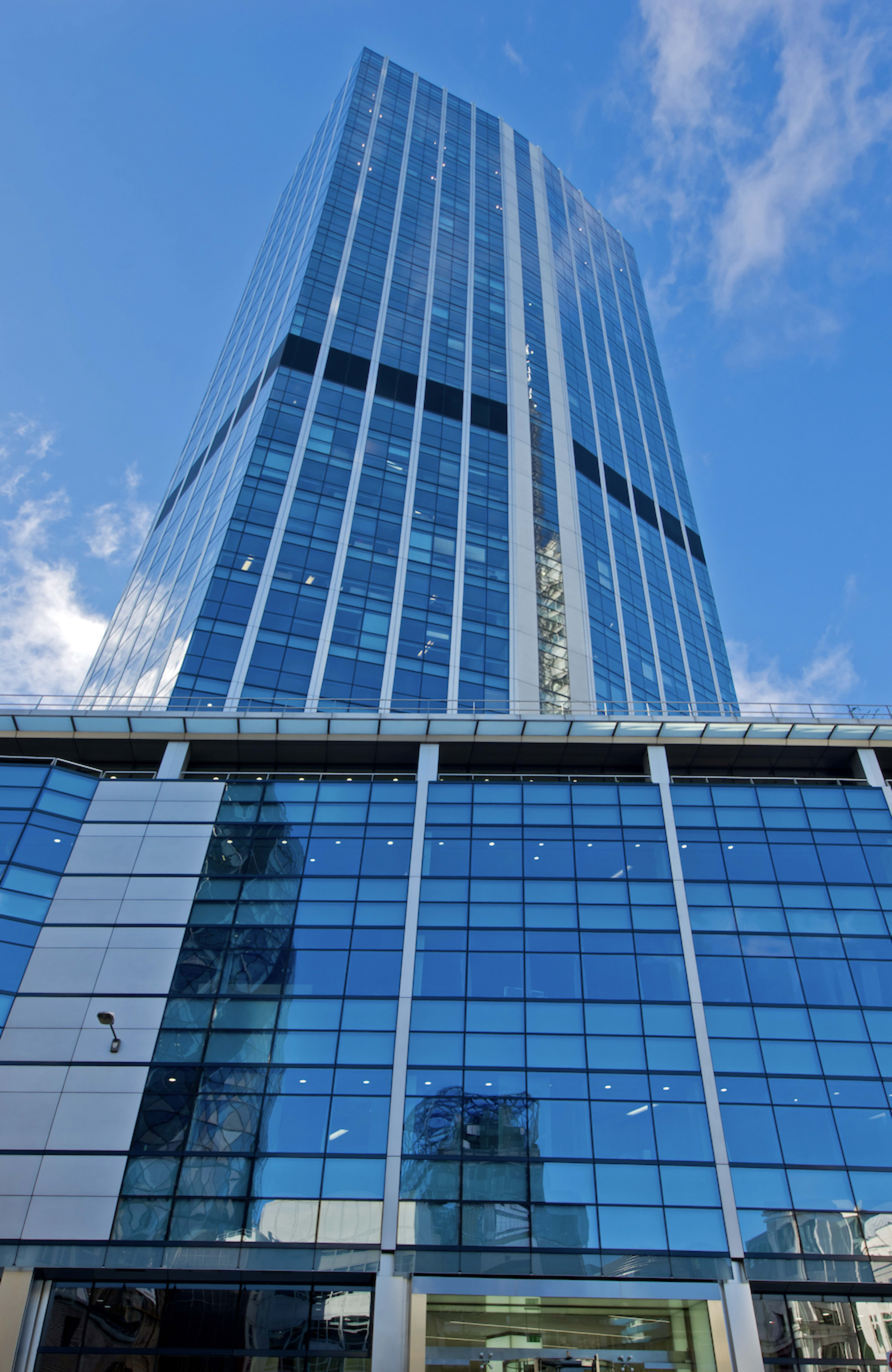A tall, modern glass skyscraper reflects the sky and surrounding buildings, with a clear blue backdrop.