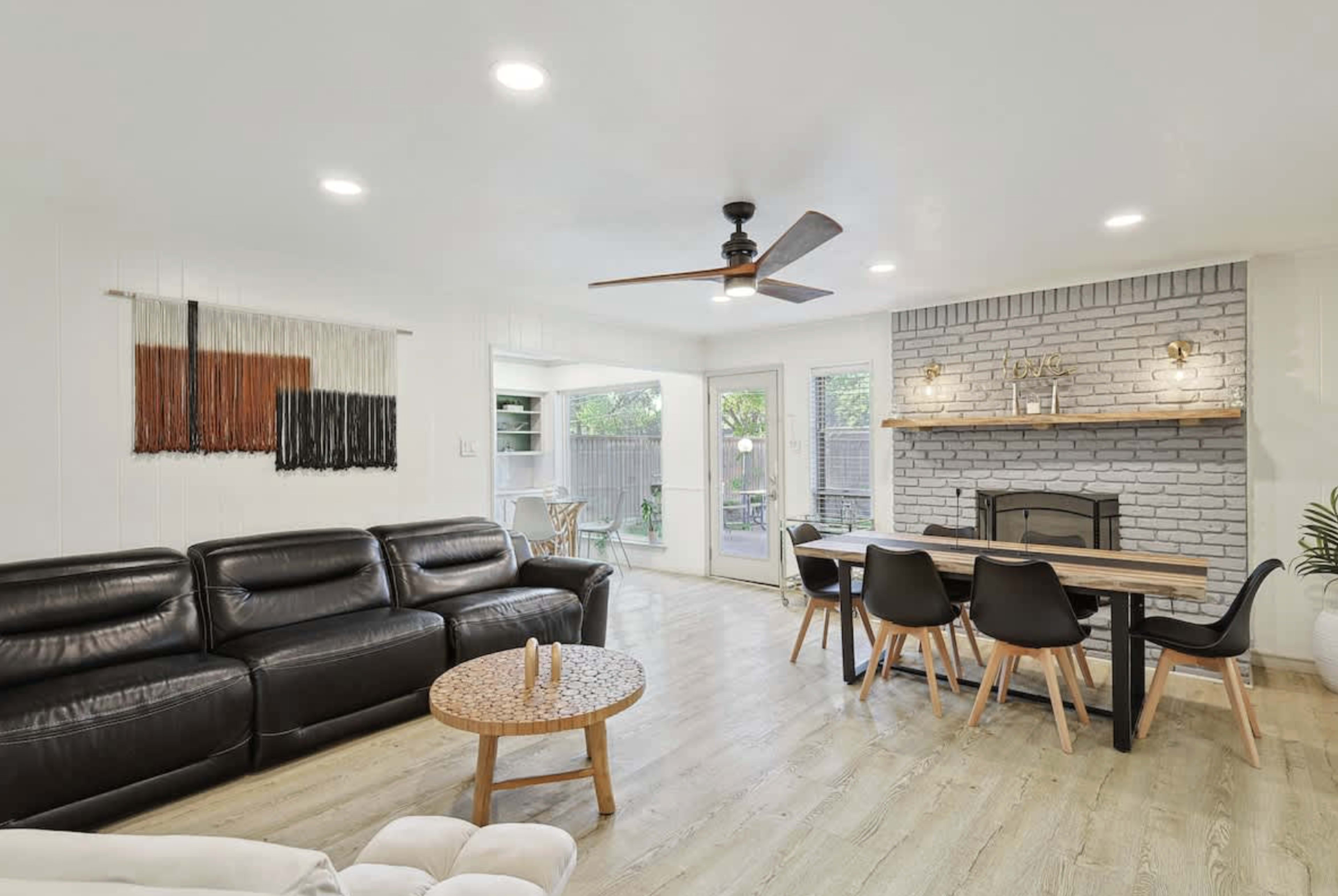 The image shows a modern living room with a black leather sofa, a wooden dining table, a fireplace with a brick surround, and sliding glass doors leading to an outdoor space.