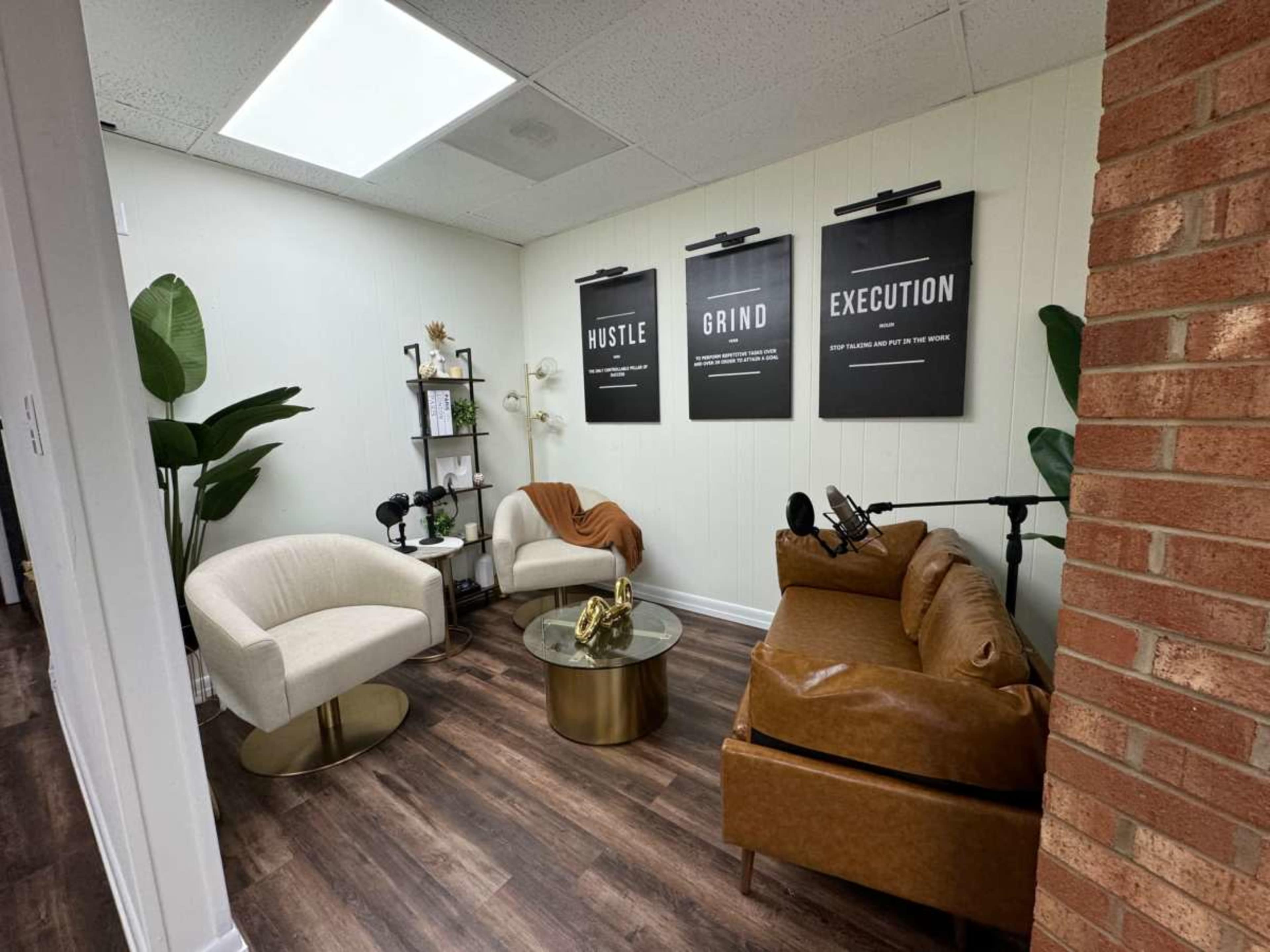 The image shows a small office or meeting space with two chairs, a brown couch, a coffee table, and motivational posters on the wall, surrounded by plants.