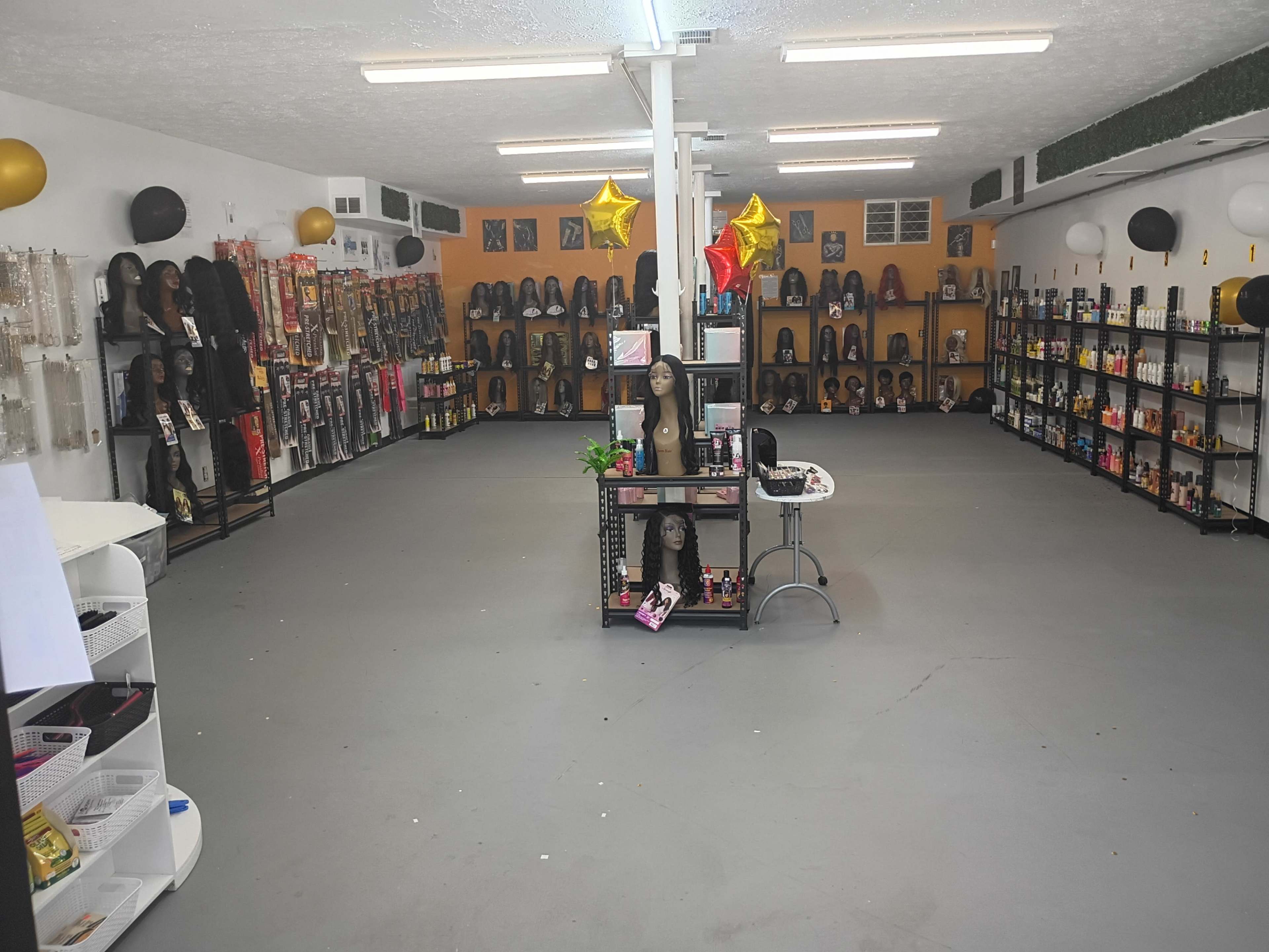 The image shows a retail space filled with displays of wigs and beauty products, with shelves lining the walls and a central table showcasing items.