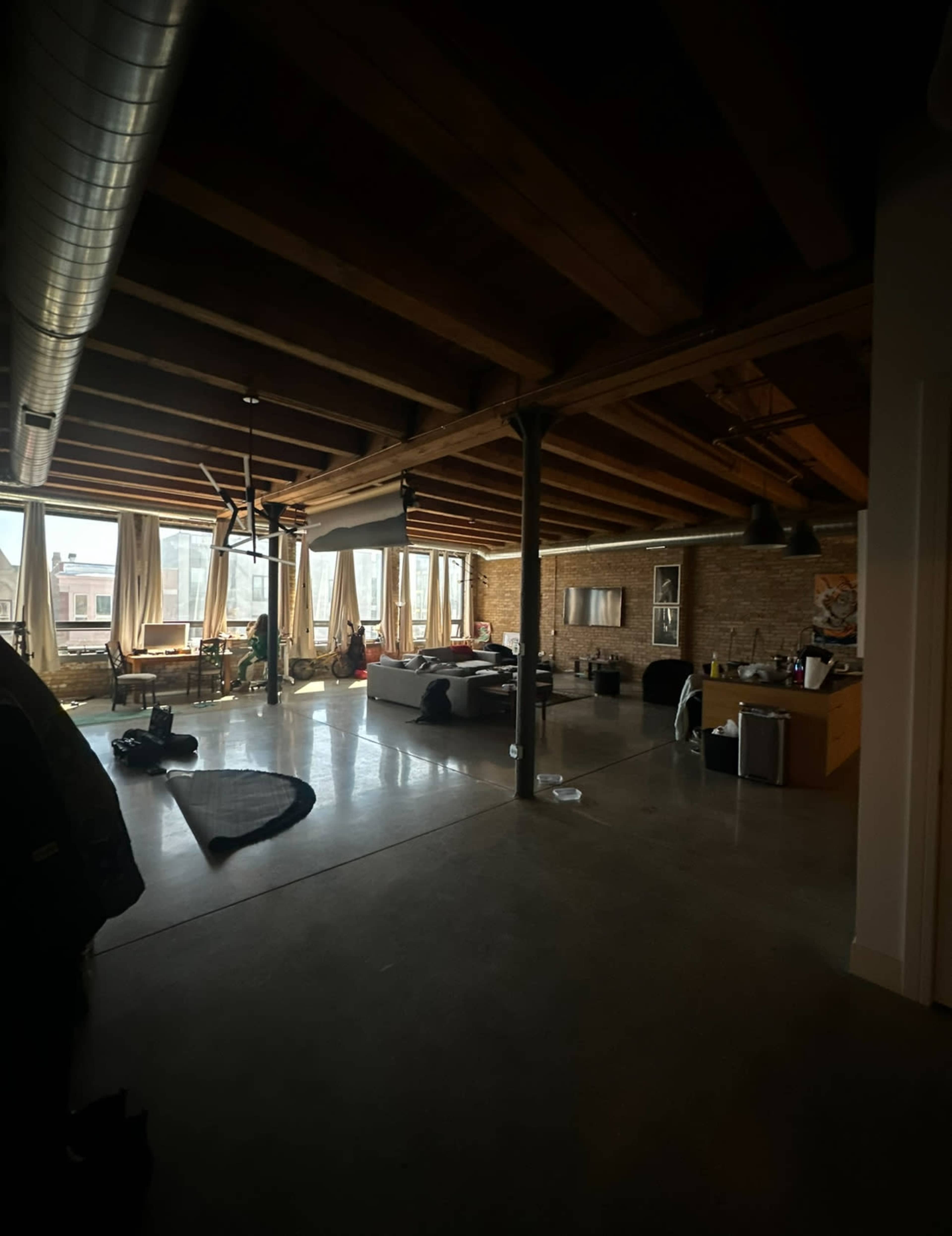 A spacious loft apartment with exposed wooden beams, large windows, and a mix of furniture scattered throughout.