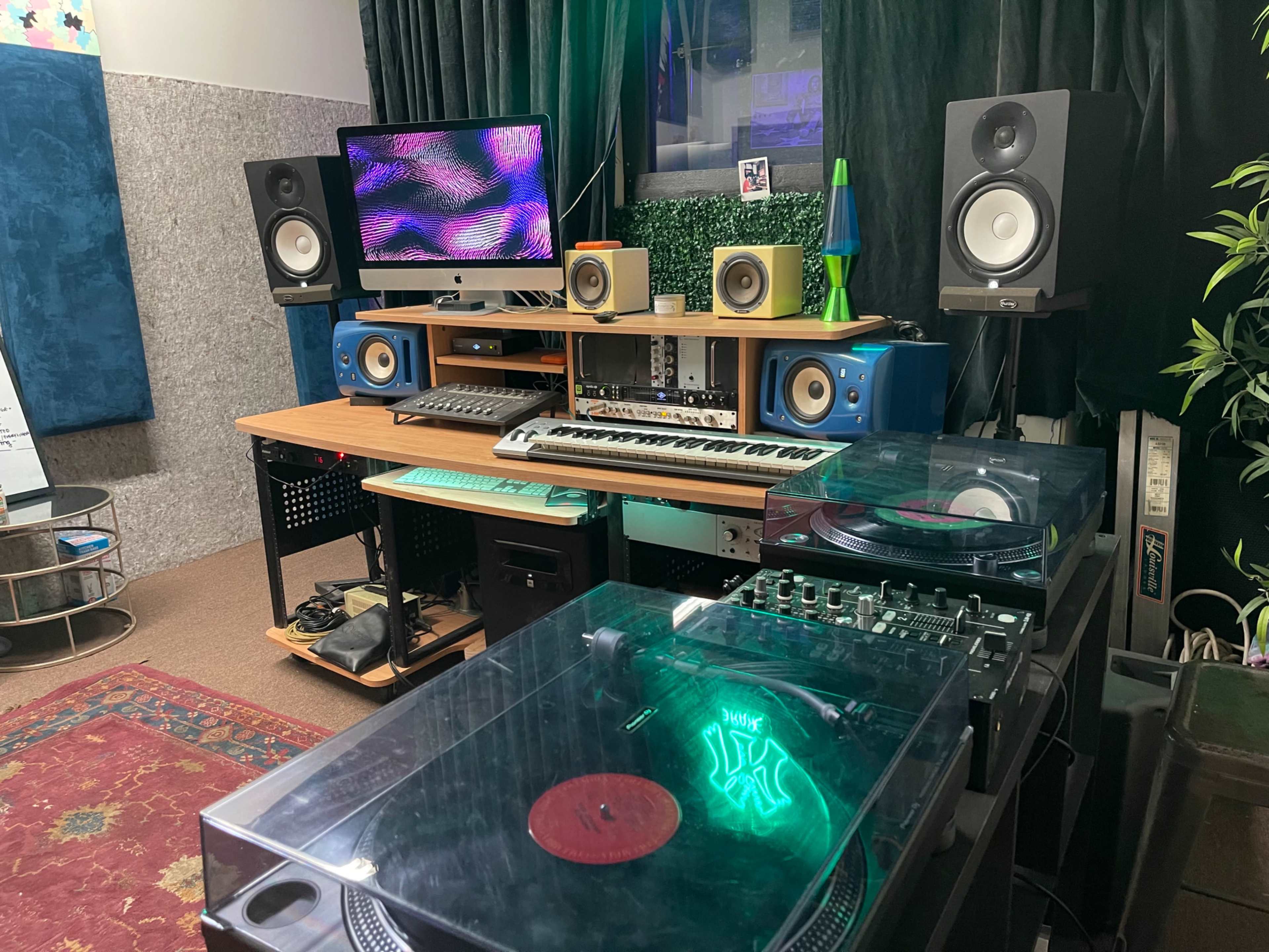 A music studio setup featuring vinyl turntables, a keyboard, speakers, and a monitor displaying colorful visuals.