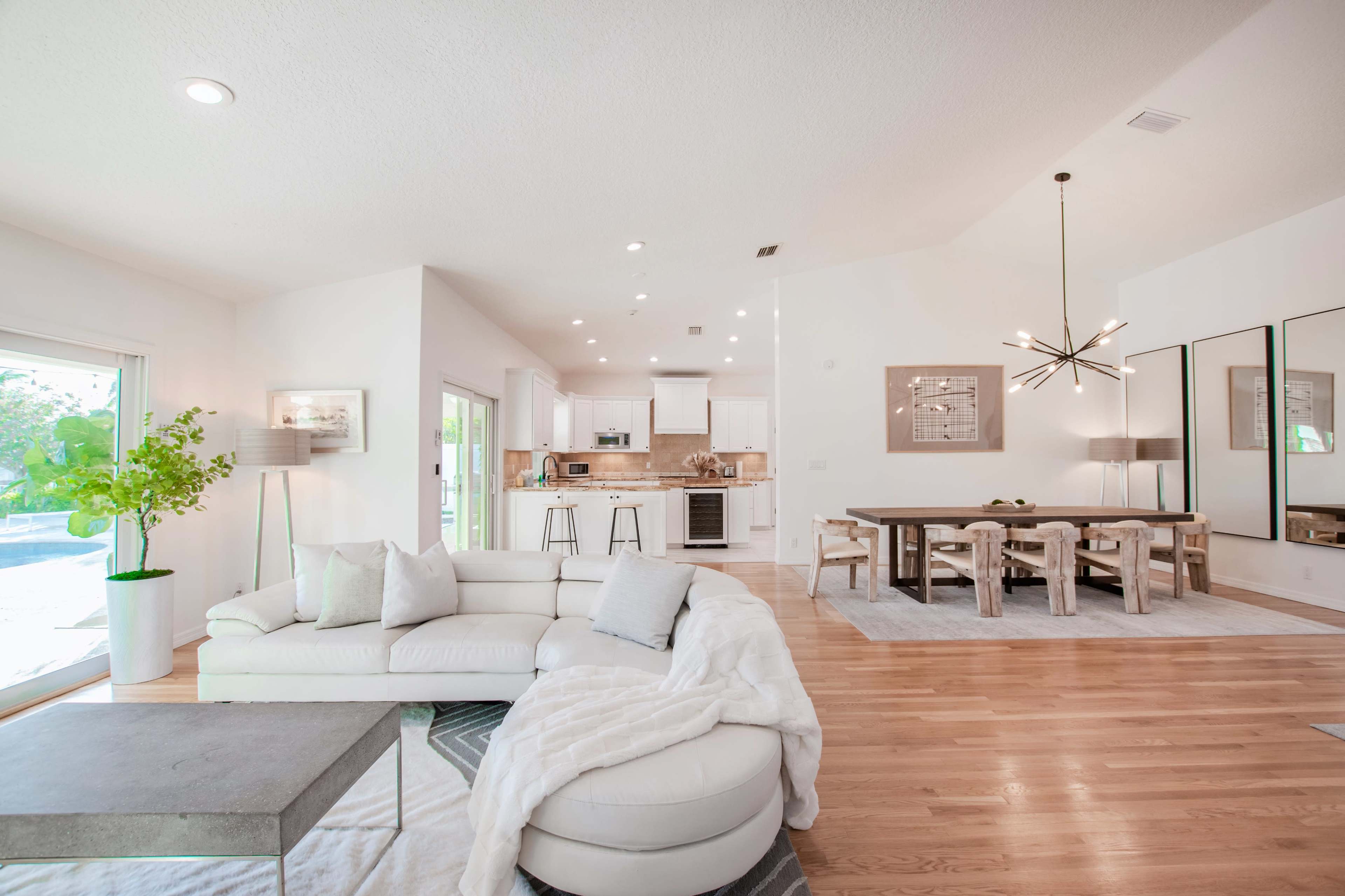 The image shows a modern living space with a white sectional sofa, a dining area featuring a large table and upholstered chairs, and a bright kitchen visible in the background.