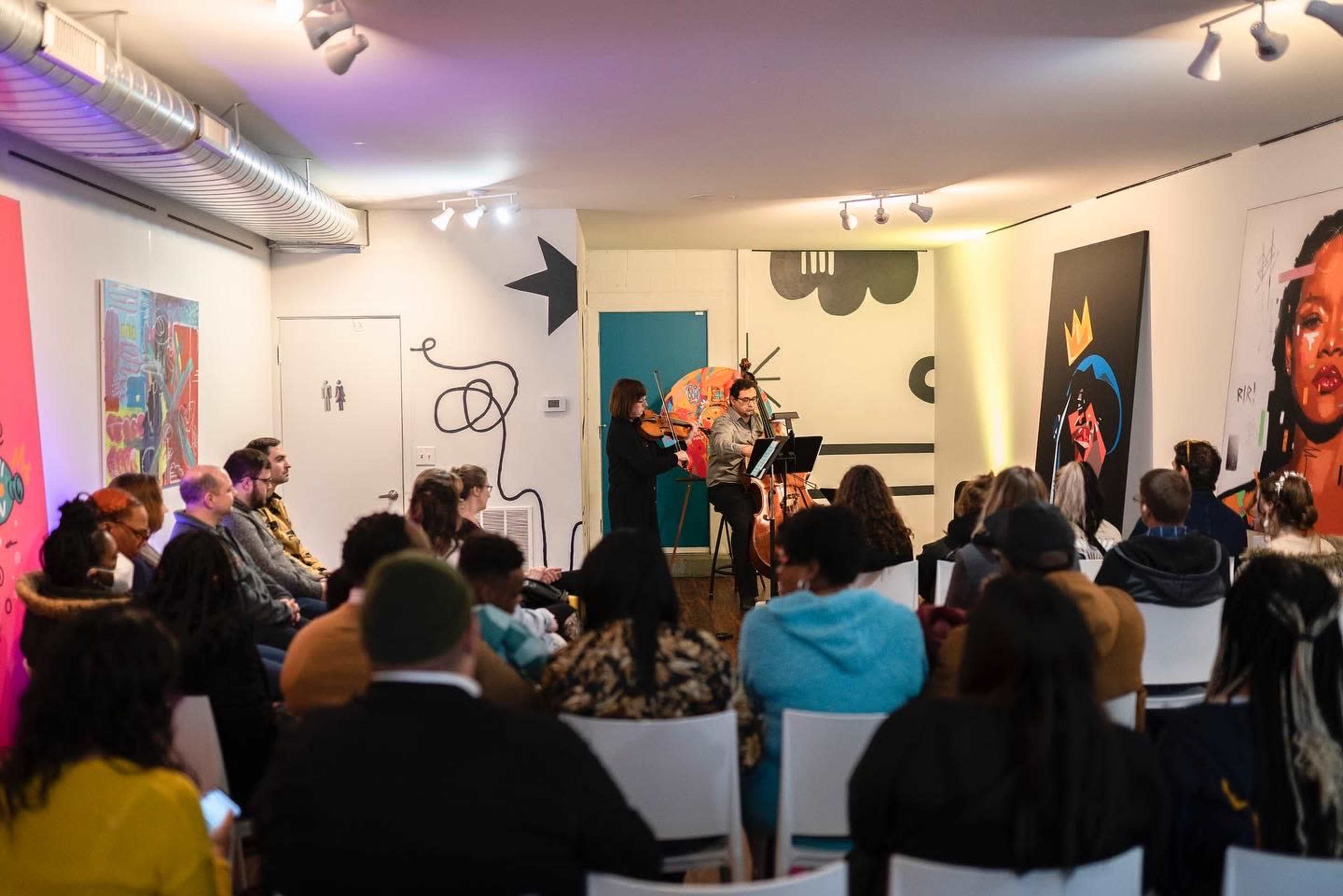 A small audience watches a music performance in a gallery featuring vibrant artwork on the walls.