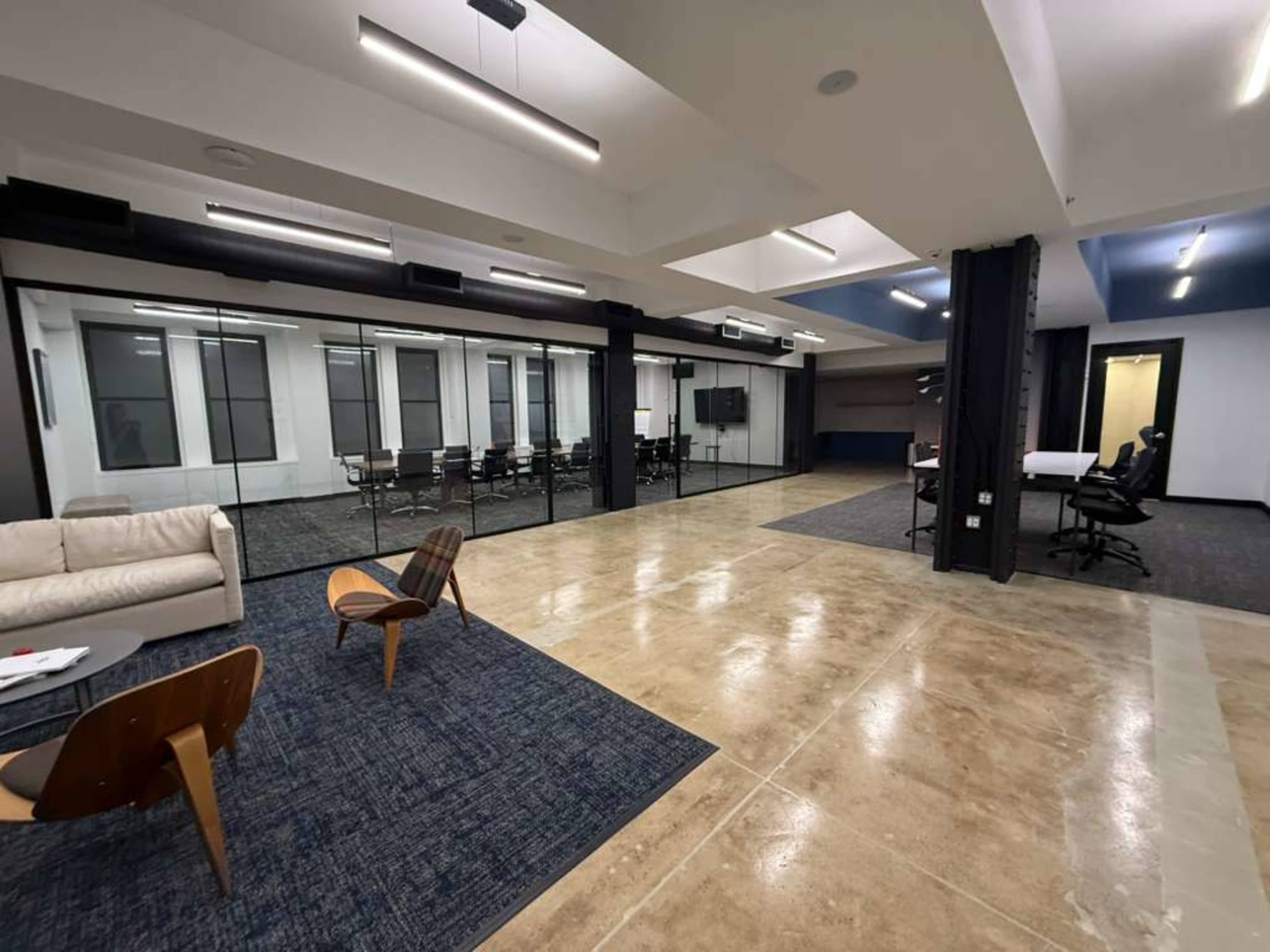 The image shows a modern office space with a seating area, glass-walled conference room, and workstations.