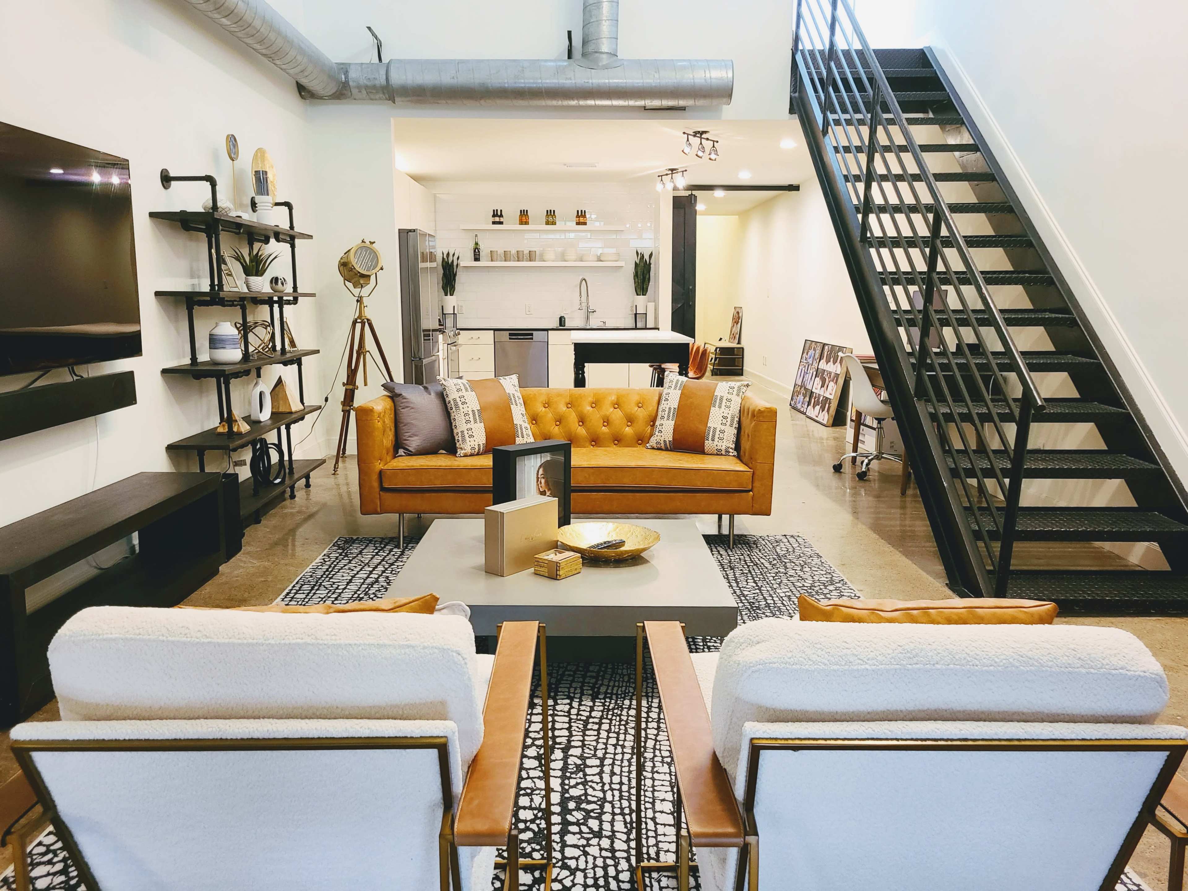 A modern living room with a brown tufted sofa, two white armchairs, a coffee table, and a staircase leading to an upper level, complemented by sleek shelves and a large television.