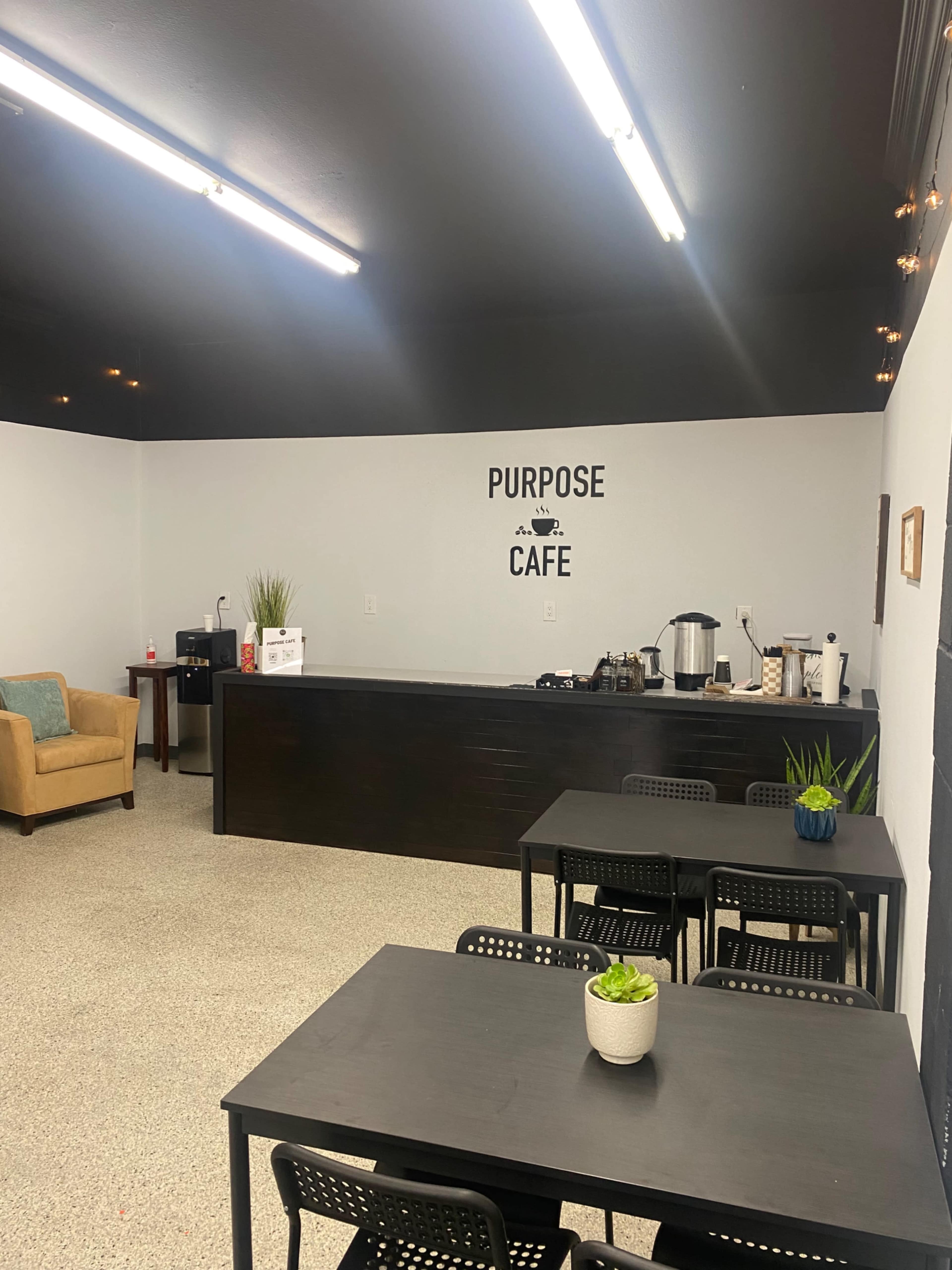 A small cafe interior with a counter, several tables, and a wall that reads "PURPOSE CAFE."