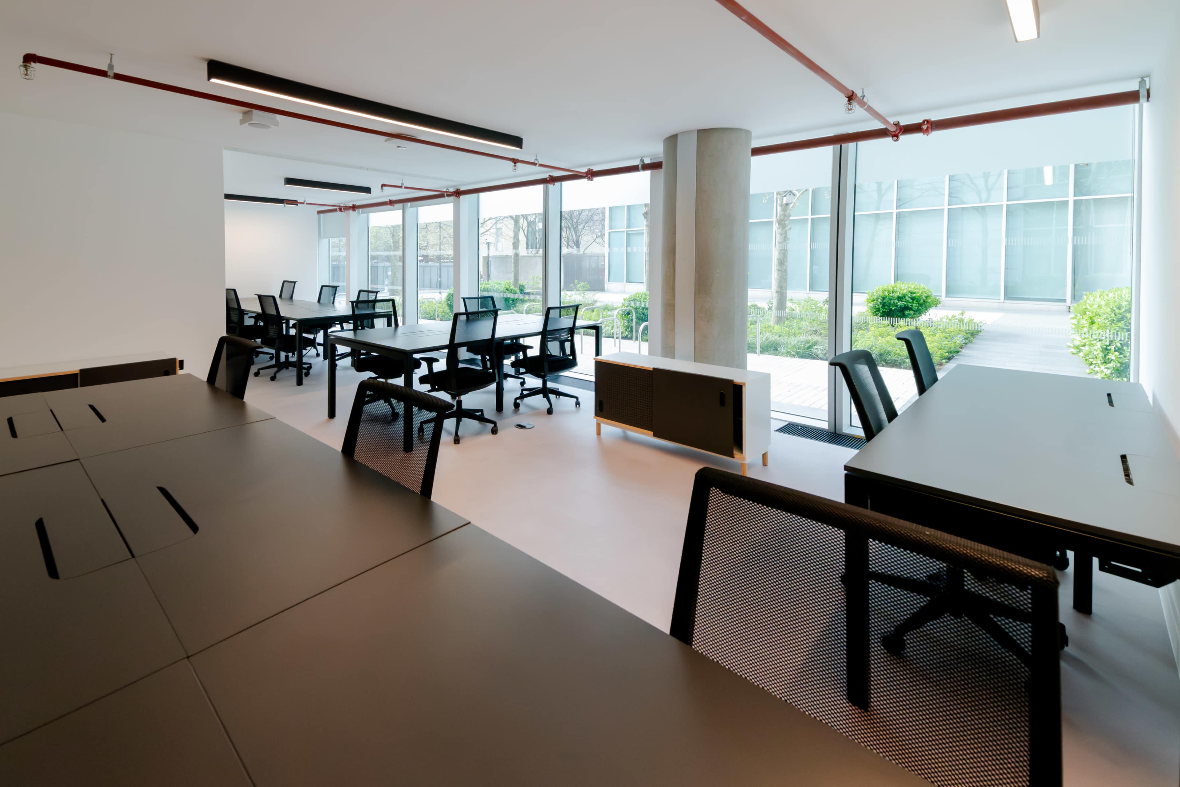 The image shows a modern office space with multiple desks and chairs arranged in a professional layout, featuring large windows that allow natural light to enter.