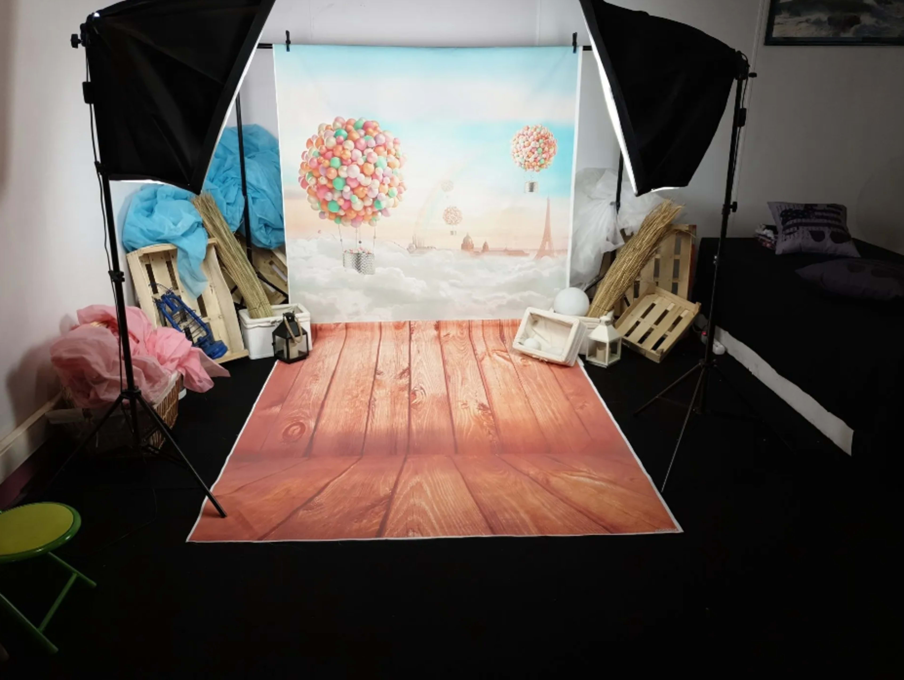 A photo studio setup features a backdrop of colorful balloons floating over a cityscape, with wooden floor props and studio lighting equipment.