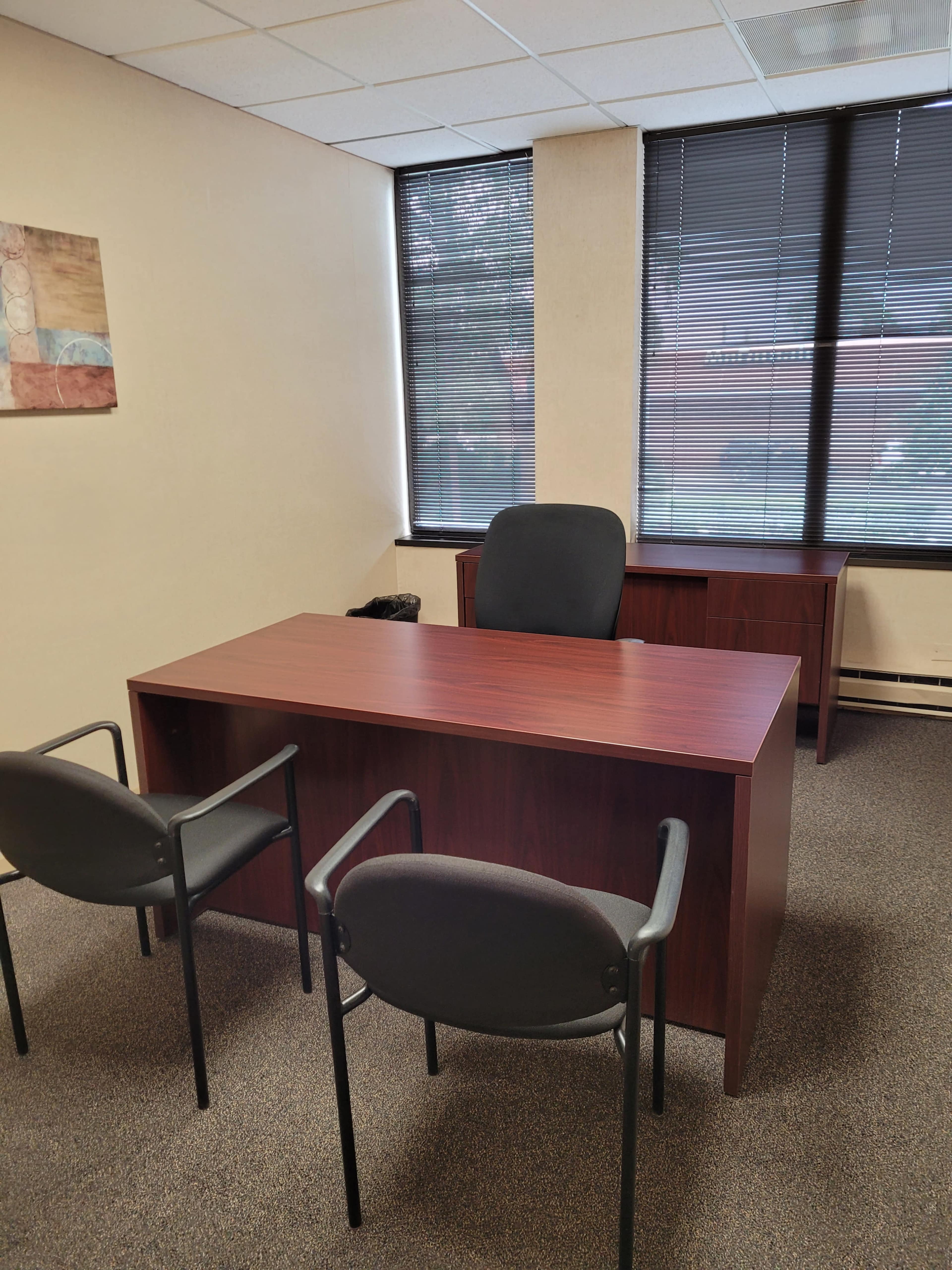 A small office with a wooden desk, two black chairs, and a larger desk against the wall, all surrounded by windows with blinds.