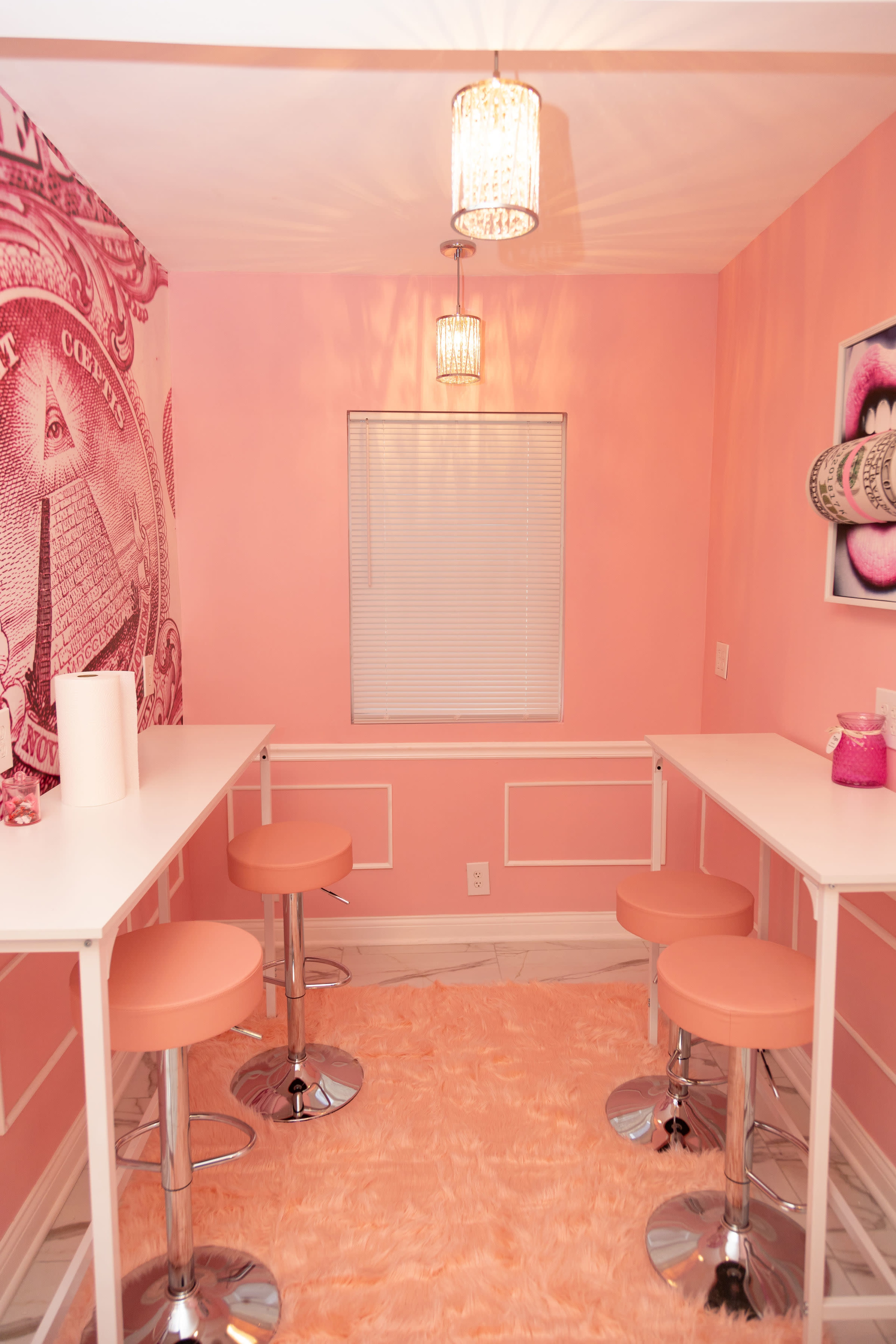 A small room with pink walls, a patterned wall mural, two white tables, pink bar stools, and a light-colored rug.