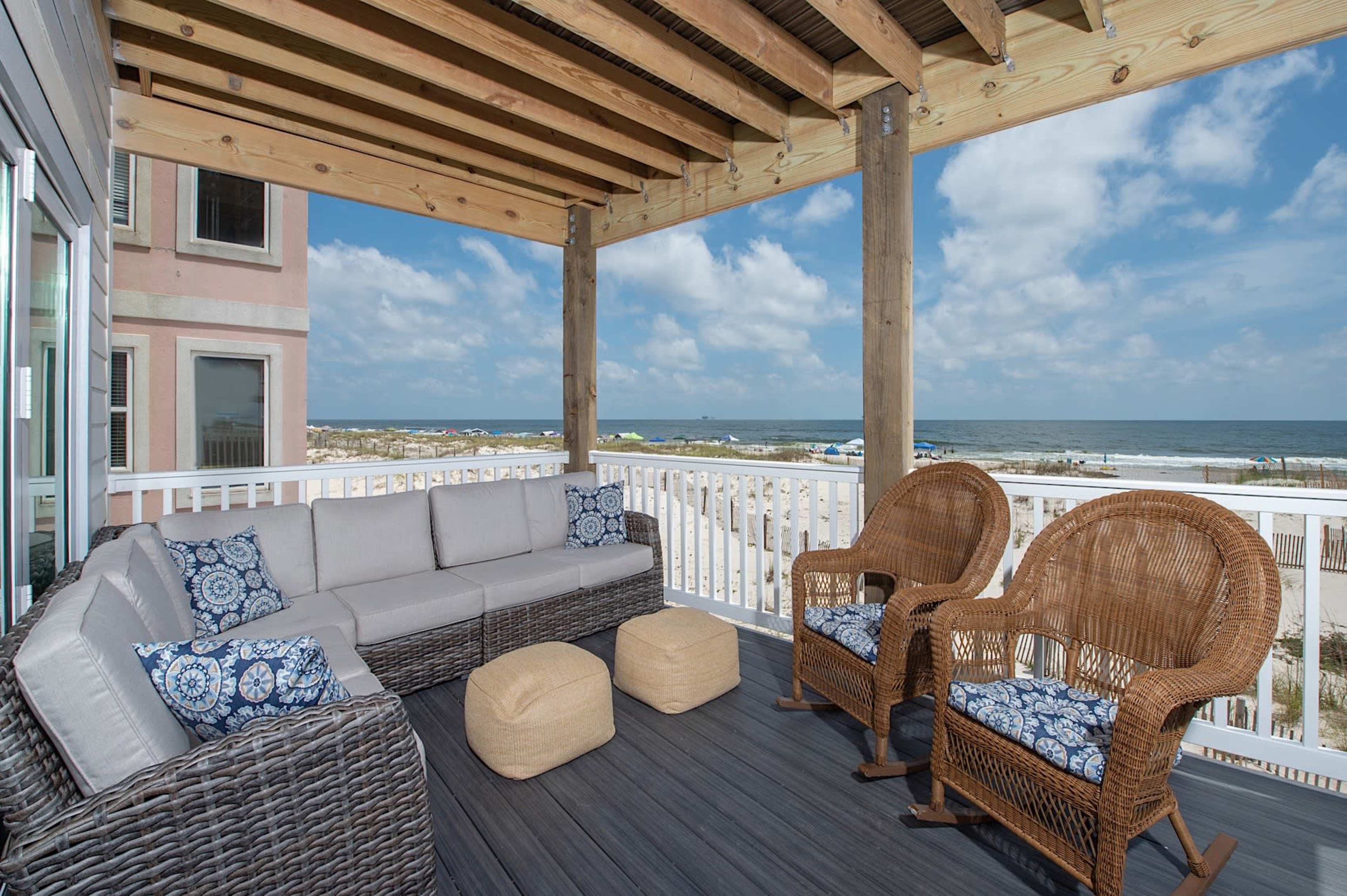 A covered porch with wicker furniture overlooks the beach and ocean under a partly cloudy sky.