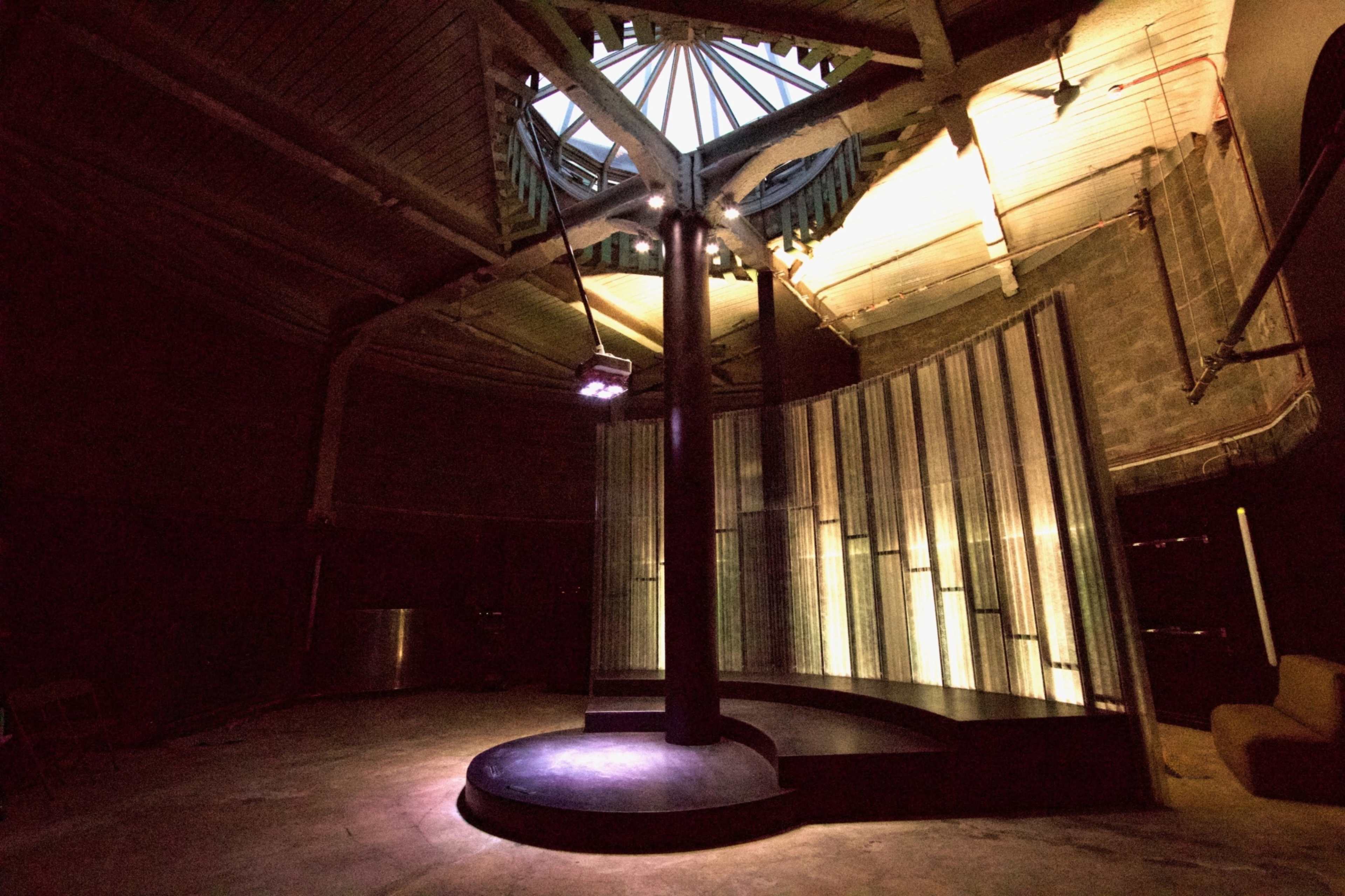 The image shows a dimly lit circular room with a central circular stage and tall, translucent panels reflecting light.