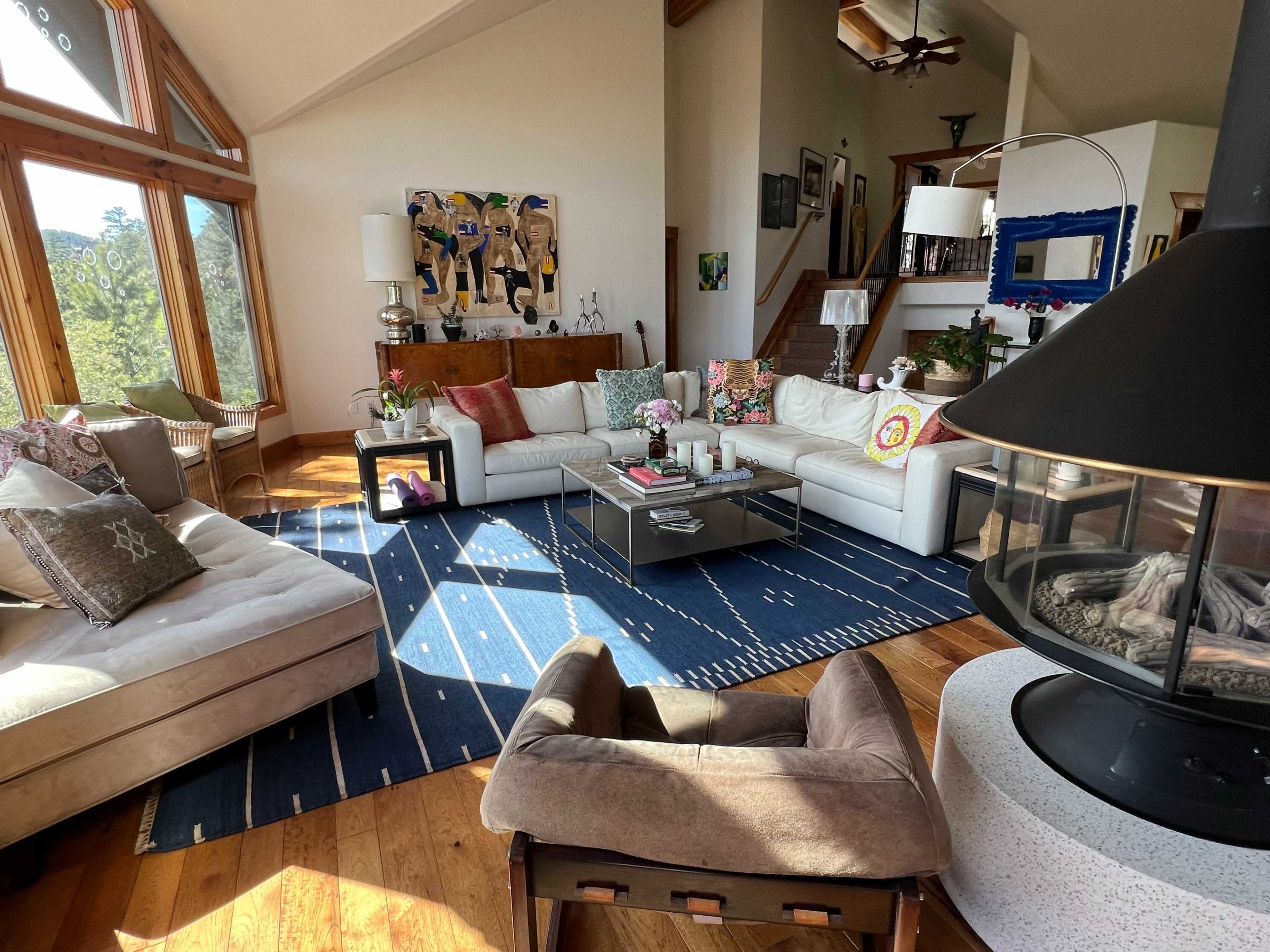 A spacious living room features large windows, modern furniture, decorative pillows, and a central fireplace.