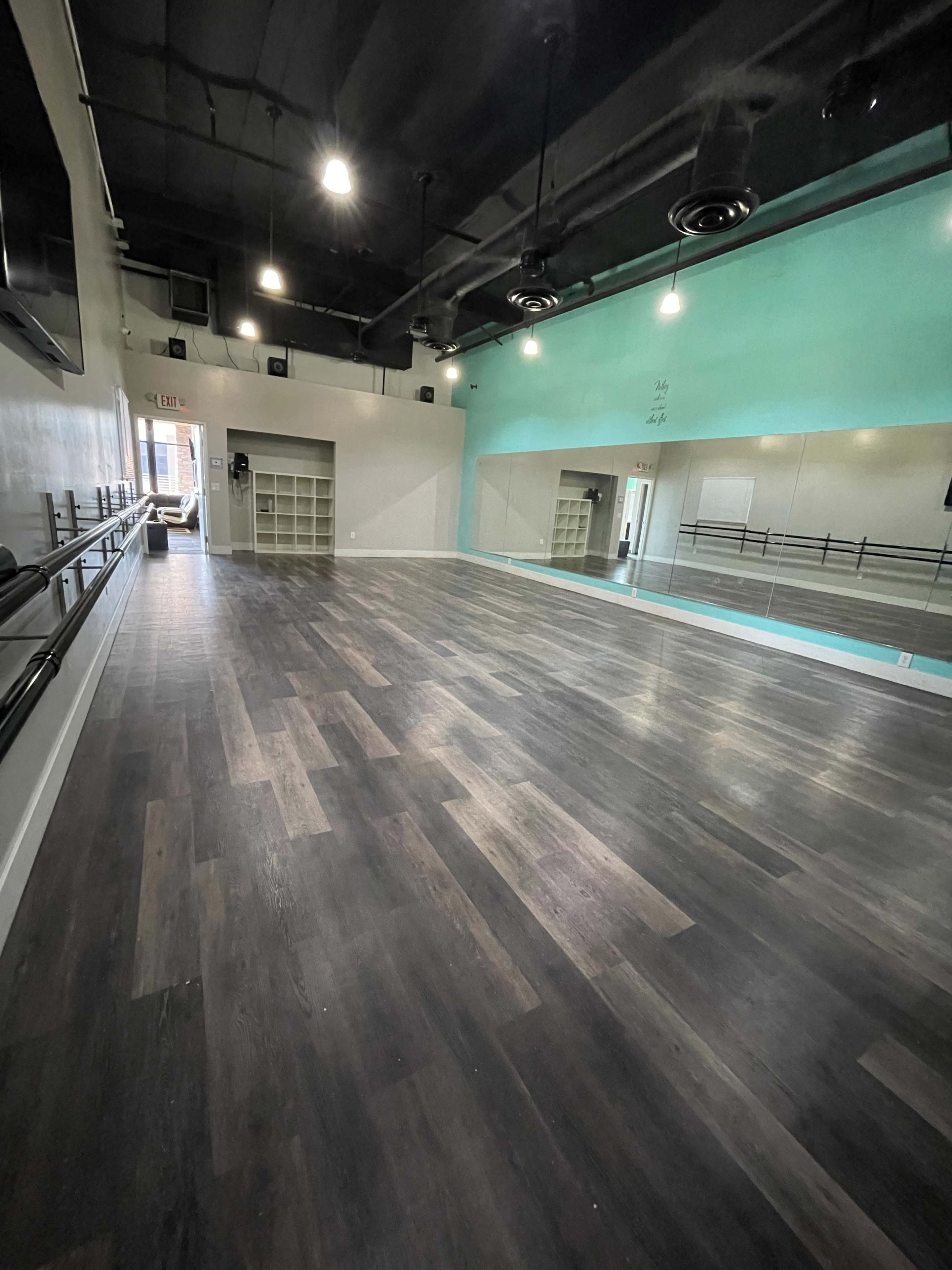 A spacious dance studio featuring a mirrored wall, wooden flooring, and bright overhead lights.