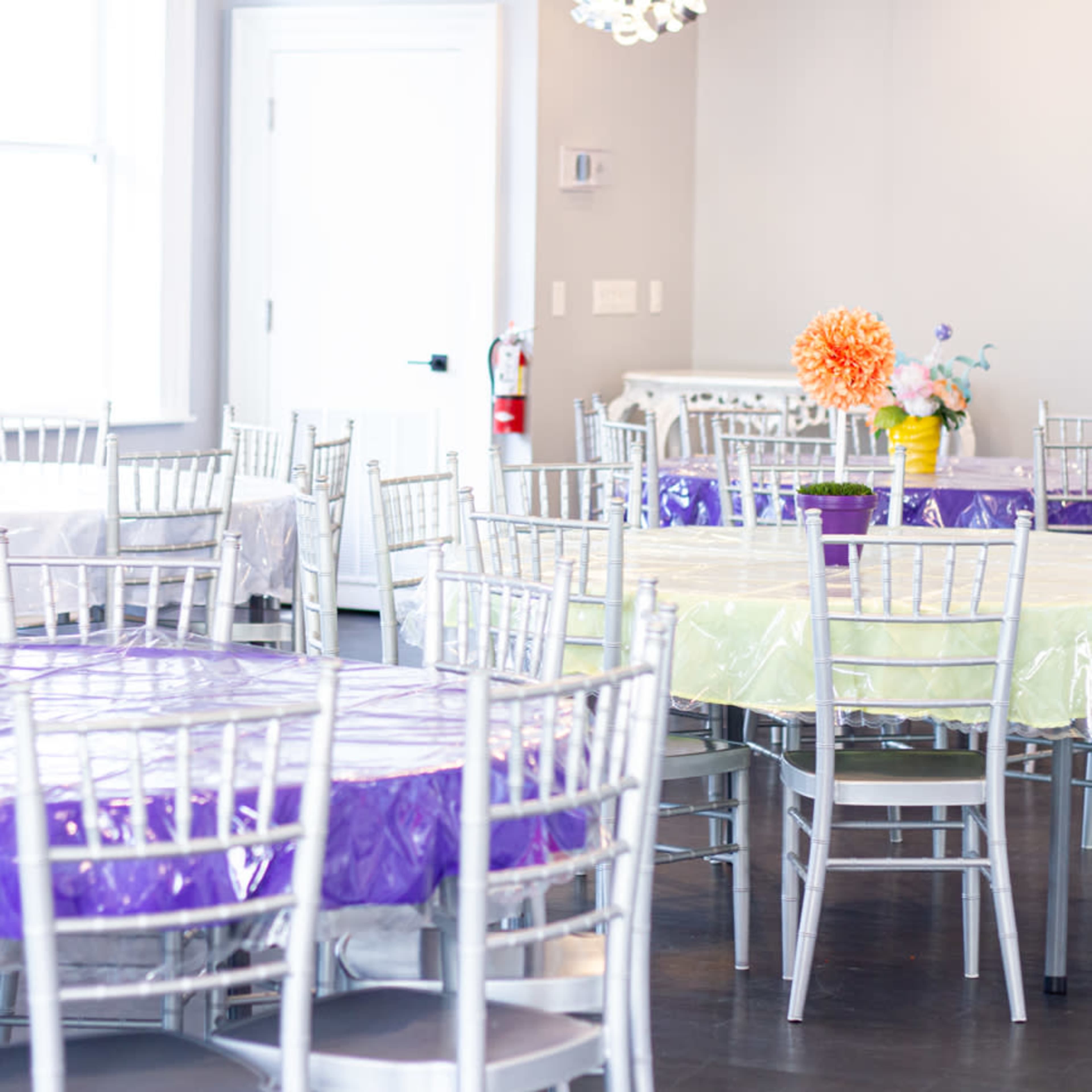 The image shows a spacious event room with multiple tables covered in various colored tablecloths and silver chairs arranged throughout the area.